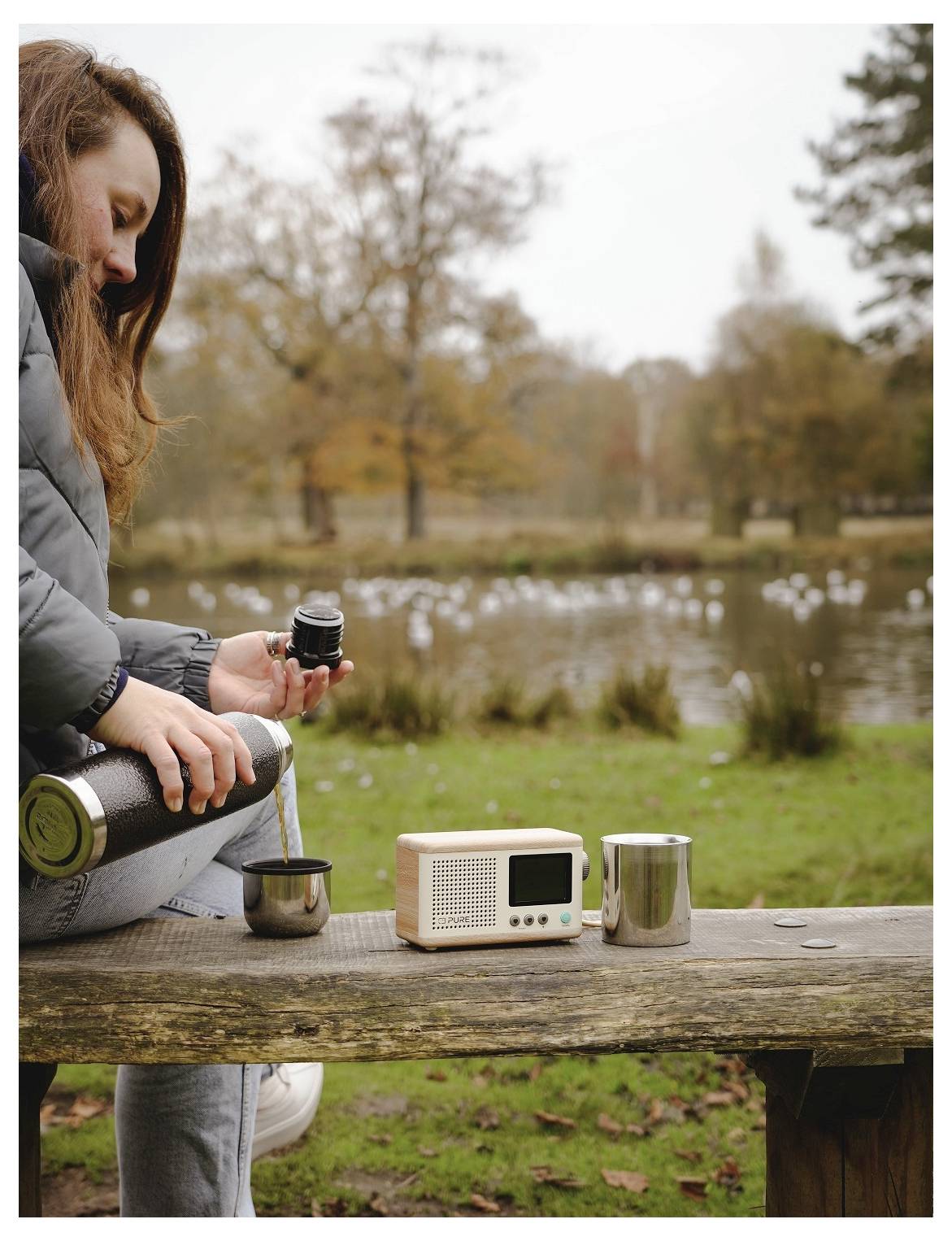 Eine Person gießt ein Getränk aus einem Thermobecher in eine Tasse neben einem Teich. Ein Radio und eine weitere Tasse liegen auf der Holzbank in der Nähe.