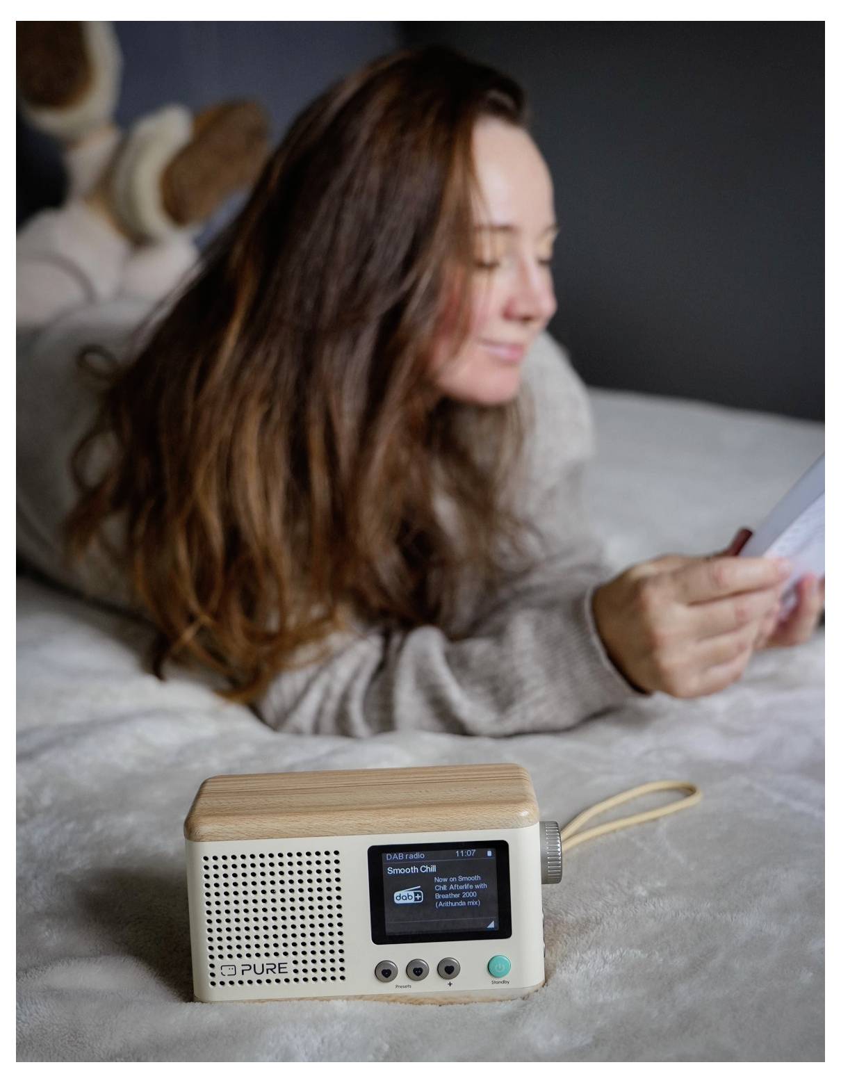 Eine Frau liegt auf einem Bett und liest eine Zeitung. Im Vordergrund befindet sich ein Radio im Retro-Stil mit einem kleinen Bildschirm und Bedienknöpfen.