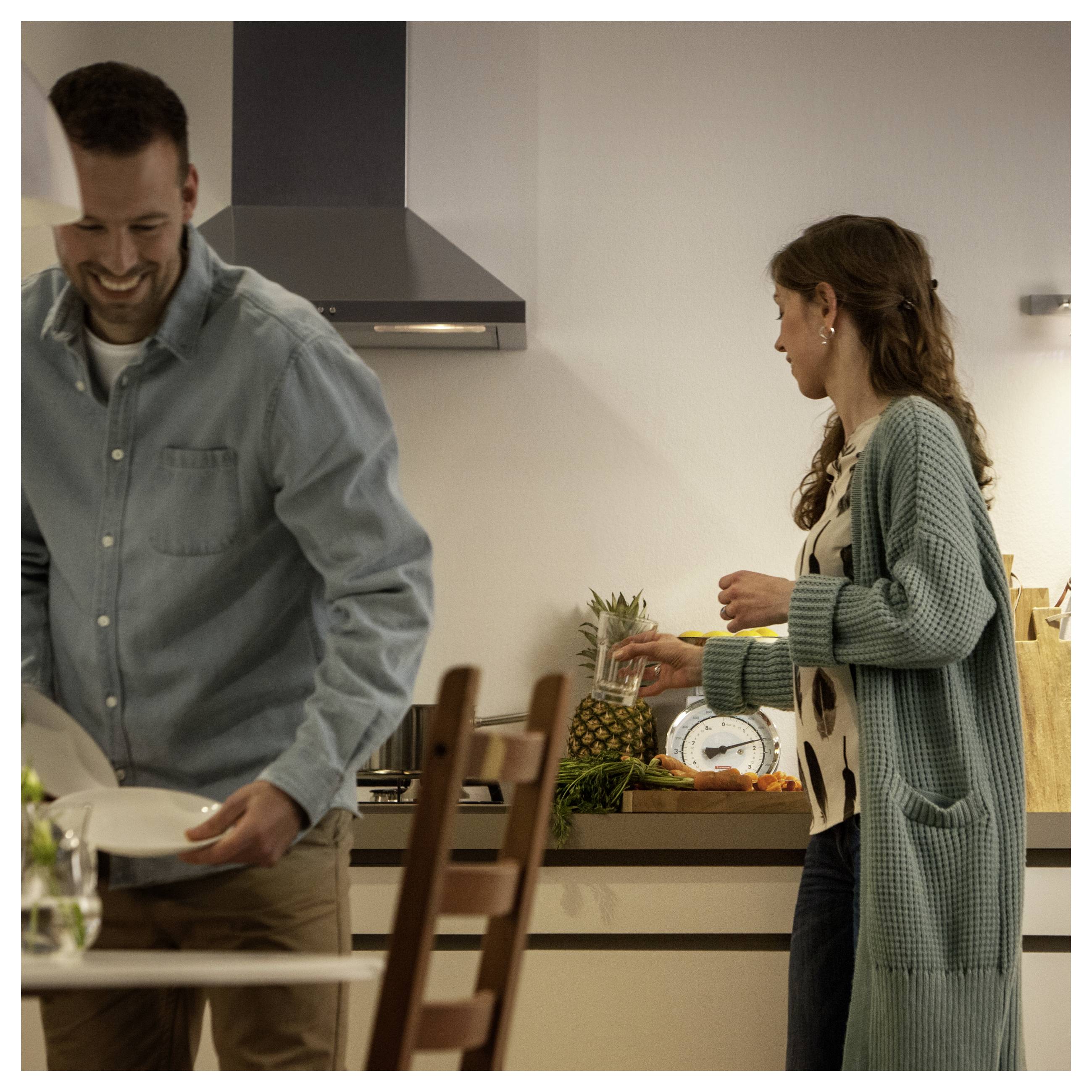 Ein Mann und eine Frau sind in einer Küche. Der Mann trägt Geschirr, während die Frau, die einen langen Cardigan trägt, in der Nähe von frischem Gemüse Essen zubereitet.