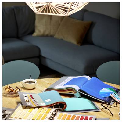 Ein Tisch mit aufgeschlagenen Farbmusterbüchern, Farbproben und einer Tasse, der vor einem grauen Sofa mit Kissen in einem dunkel beleuchteten Raum steht.