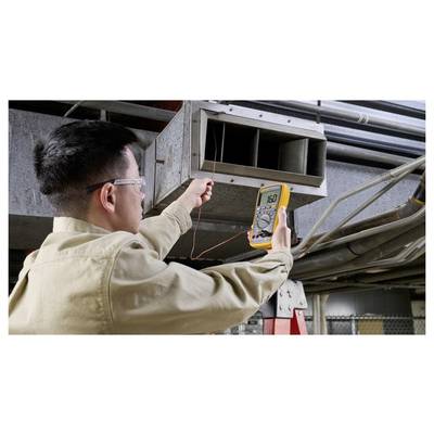 Eine Person, die mit einem digitalen Multimeter in einer industriellen Umgebung nahe einem Luftkanal die Spannung misst und dabei Arbeiten zur elektrischen Überprüfung durchführt.