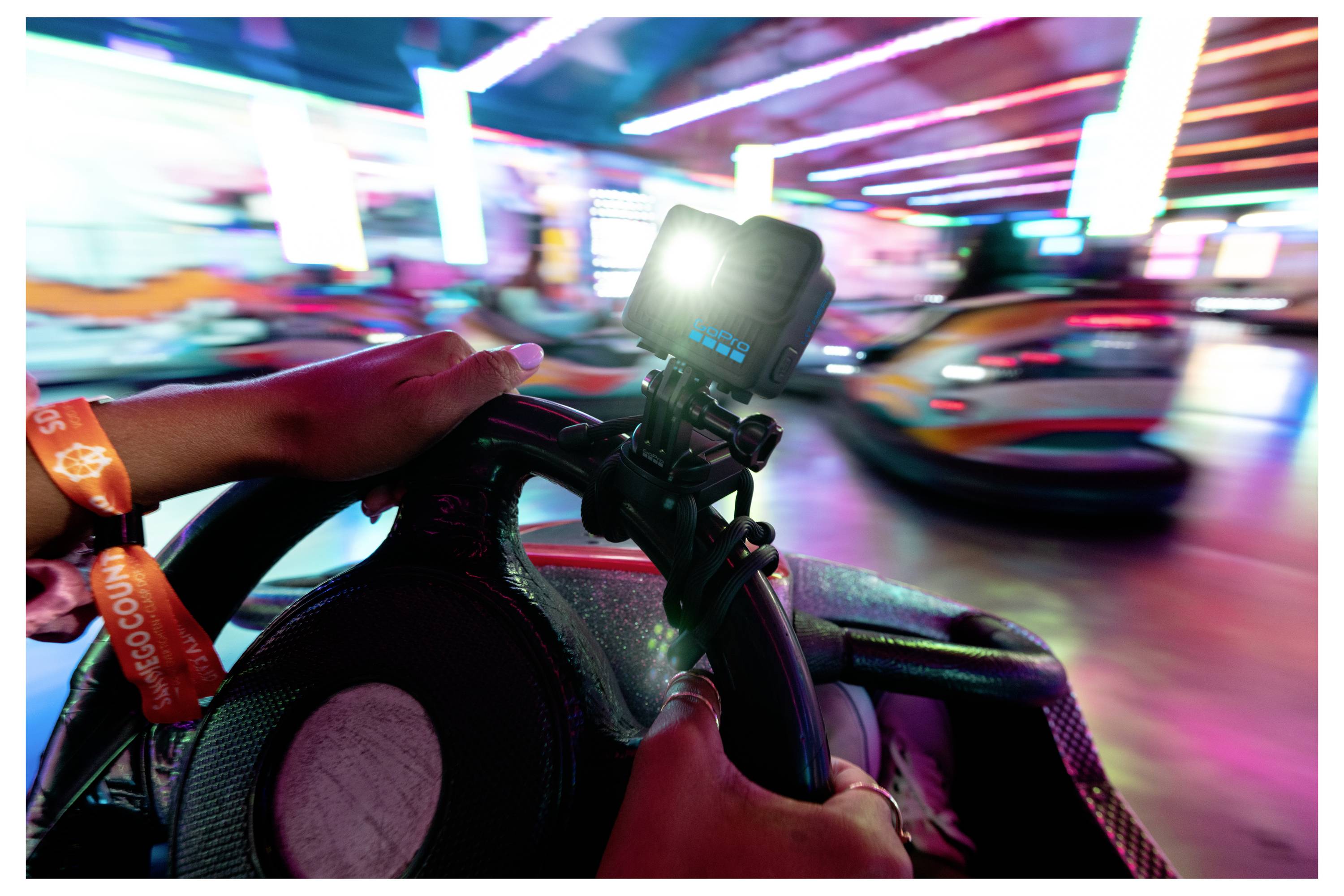 Eine Person steuert ein Autoscooter in einer Indoor-Vergnügungsanlage und hält ein Lenkrad, an dem eine Kamera montiert ist. Helle Lichter verwischen in Bewegung.