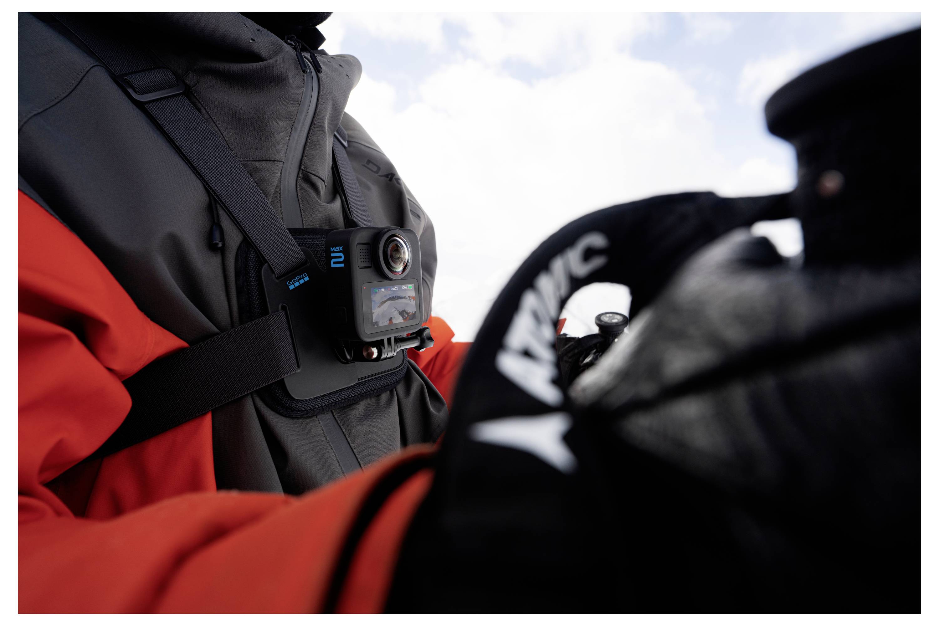 Eine Person mit einer roten und grauen Jacke, an deren Brust eine Kamera montiert ist, hält einen Gegenstand. Der Hintergrund zeigt einen bewölkten Himmel.