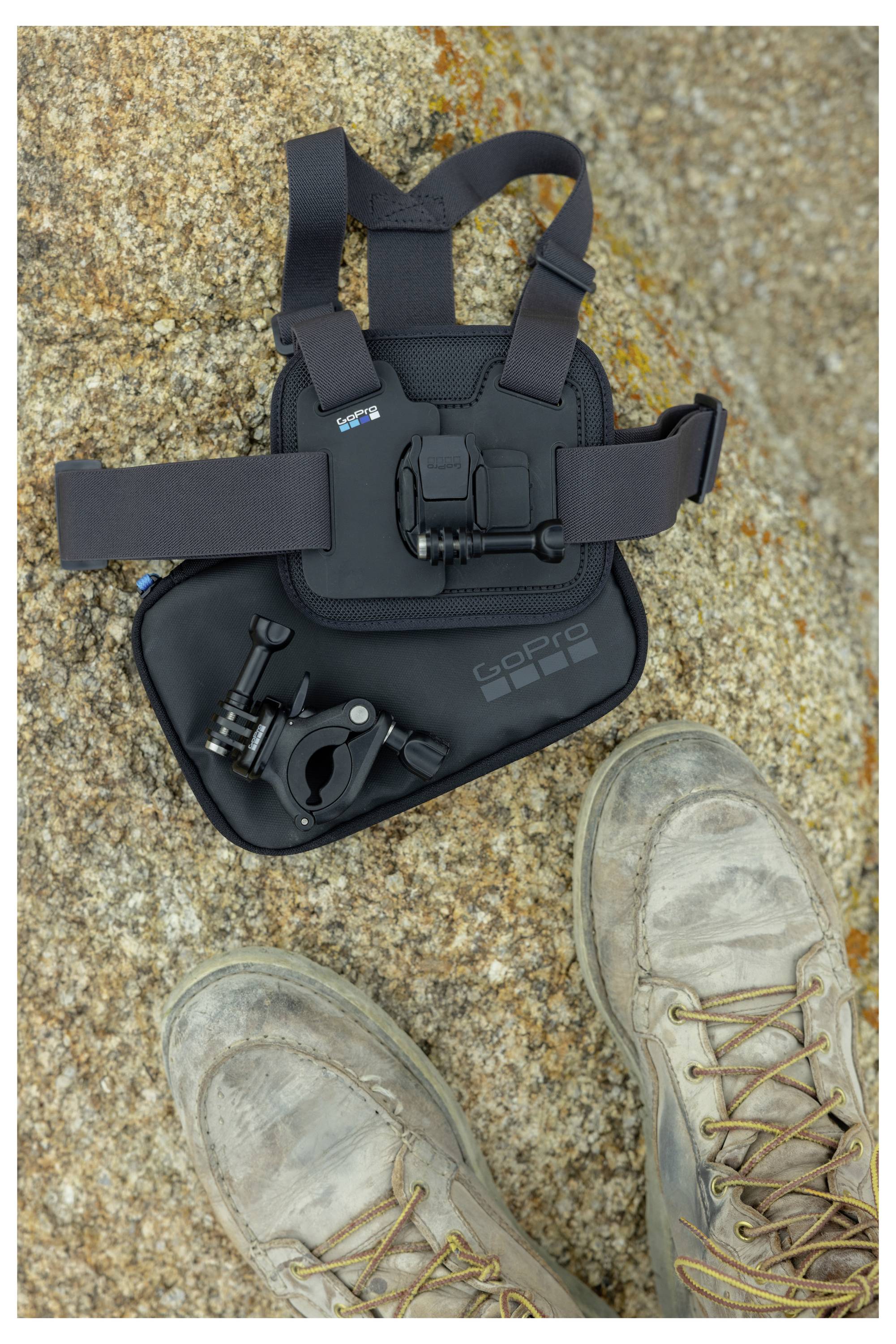 Wanderschuhe auf felsigem Gelände neben einem GoPro-Brustgurt-Geschirr und Zubehör, was eine Outdoor-Fotografie-Ausrüstung für Abenteuer andeutet.