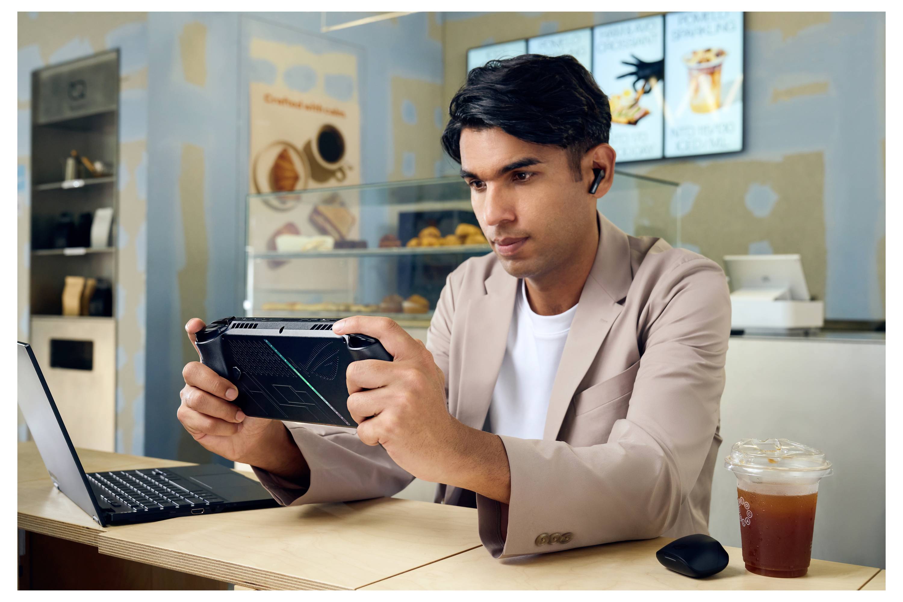 Eine Person, die an einem Cafétisch sitzt und ein tragbares Spielgerät bedient, mit einem geöffneten Laptop und einer Tasse Eiskaffee in der Nähe.