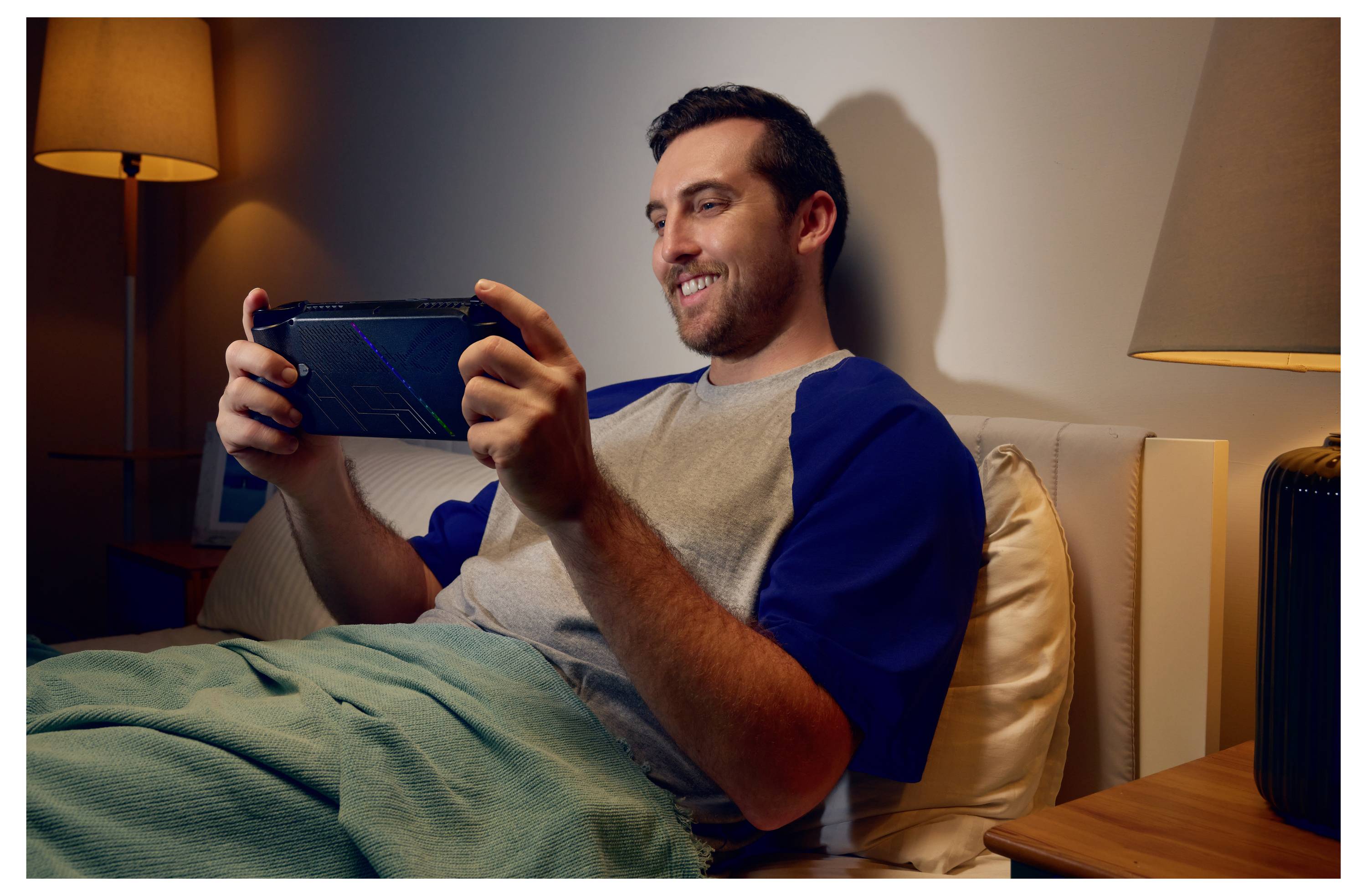 Ein Mann sitzt im Bett unter einer Decke und lächelt, während er eine tragbare Spielkonsole bedient. Eine Lampe ist daneben angezündet und schafft eine gemütliche Atmosphäre.