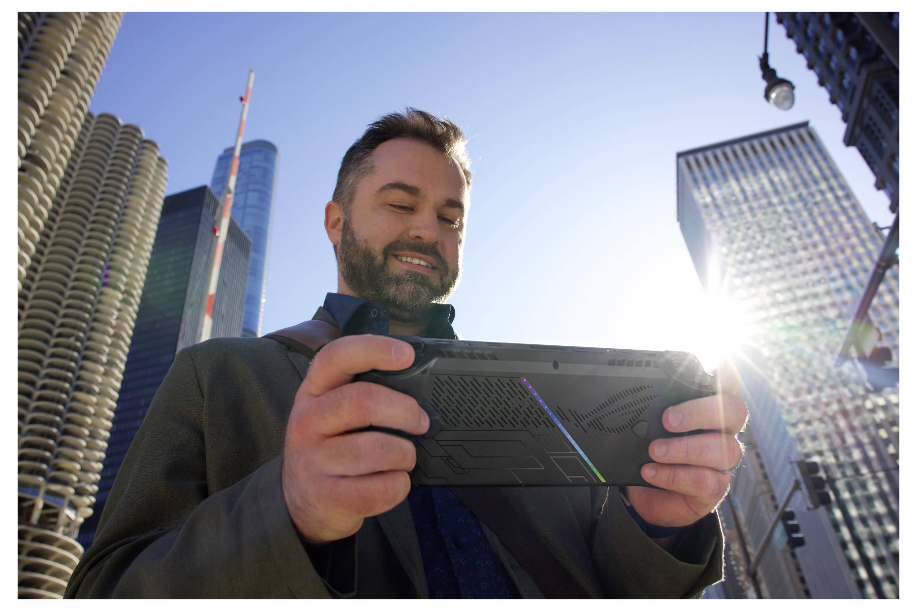 Ein Mann lächelt, während er auf einer Stadtstraße eine Spielkonsole hält. Wolkenkratzer und Sonnenlicht sind im Hintergrund.