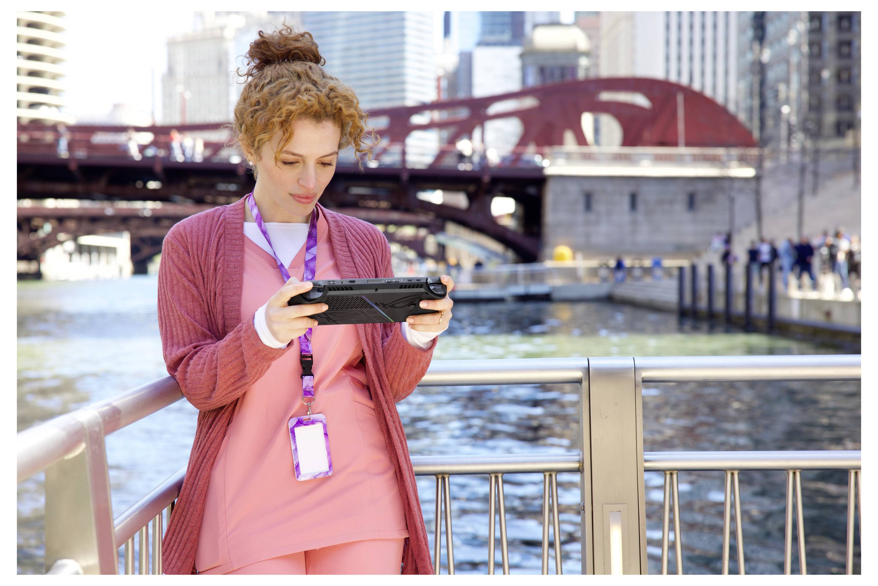 Eine Frau steht an einem Geländer in der Nähe eines Flusses und konzentriert sich auf ein Tablet-Gerät. Eine Stadtbrücke und Gebäude sind im Hintergrund zu sehen.