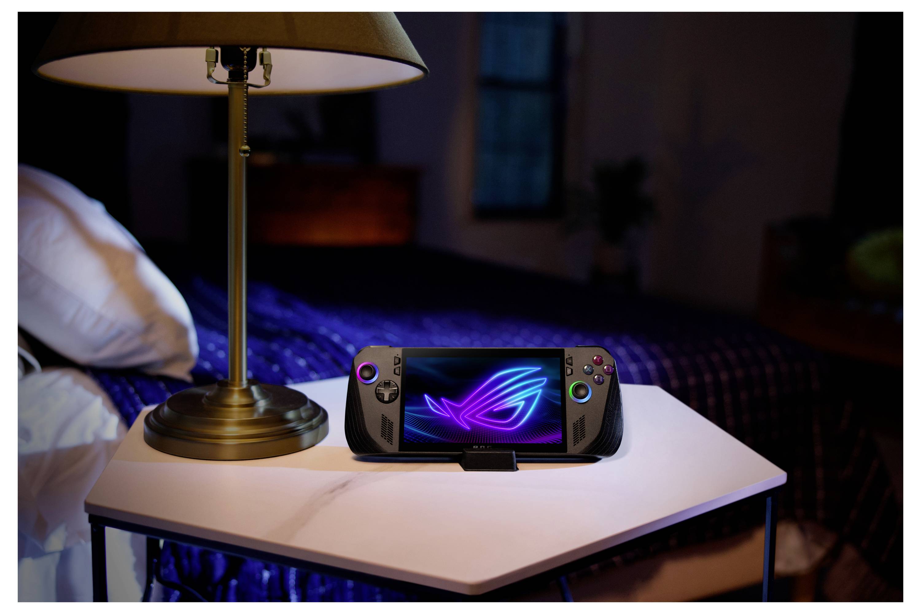 Tragbares Spielgerät mit beleuchtetem Logobilschirm auf einem Tisch neben einer Lampe, in einem dämmrig beleuchteten Schlafzimmer mit einem ordentlich gemachten Bett und einer blauen Bettdecke.