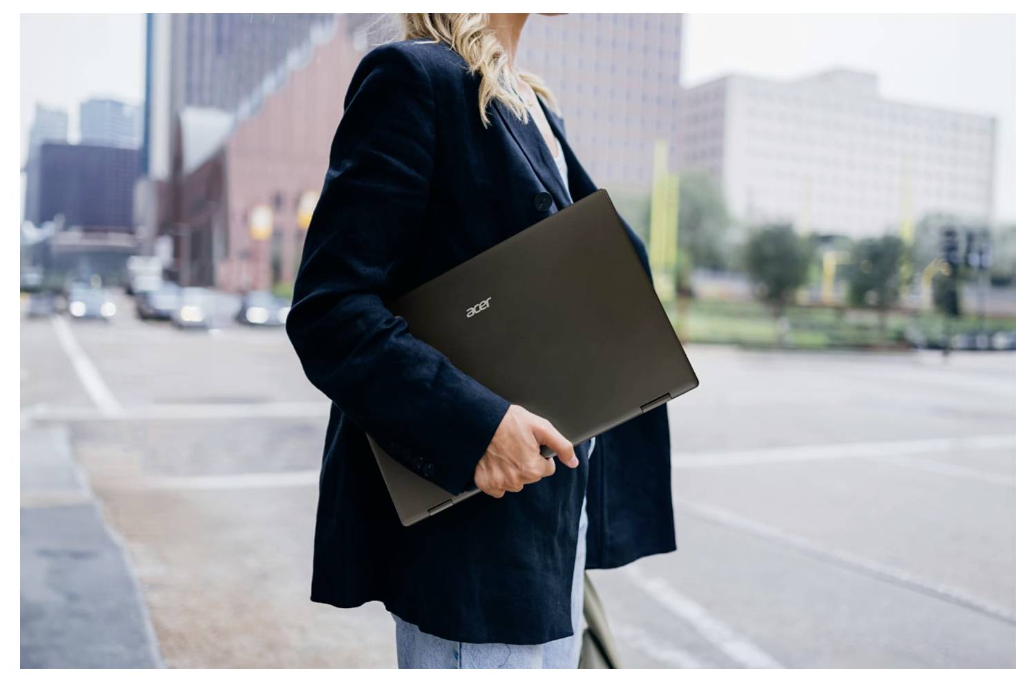 Eine Person mit einem Laptop steht an einer städtischen Kreuzung, im Hintergrund sind Stadtgebäude zu sehen, was einen Geschäfts- oder Reisekontext andeutet.