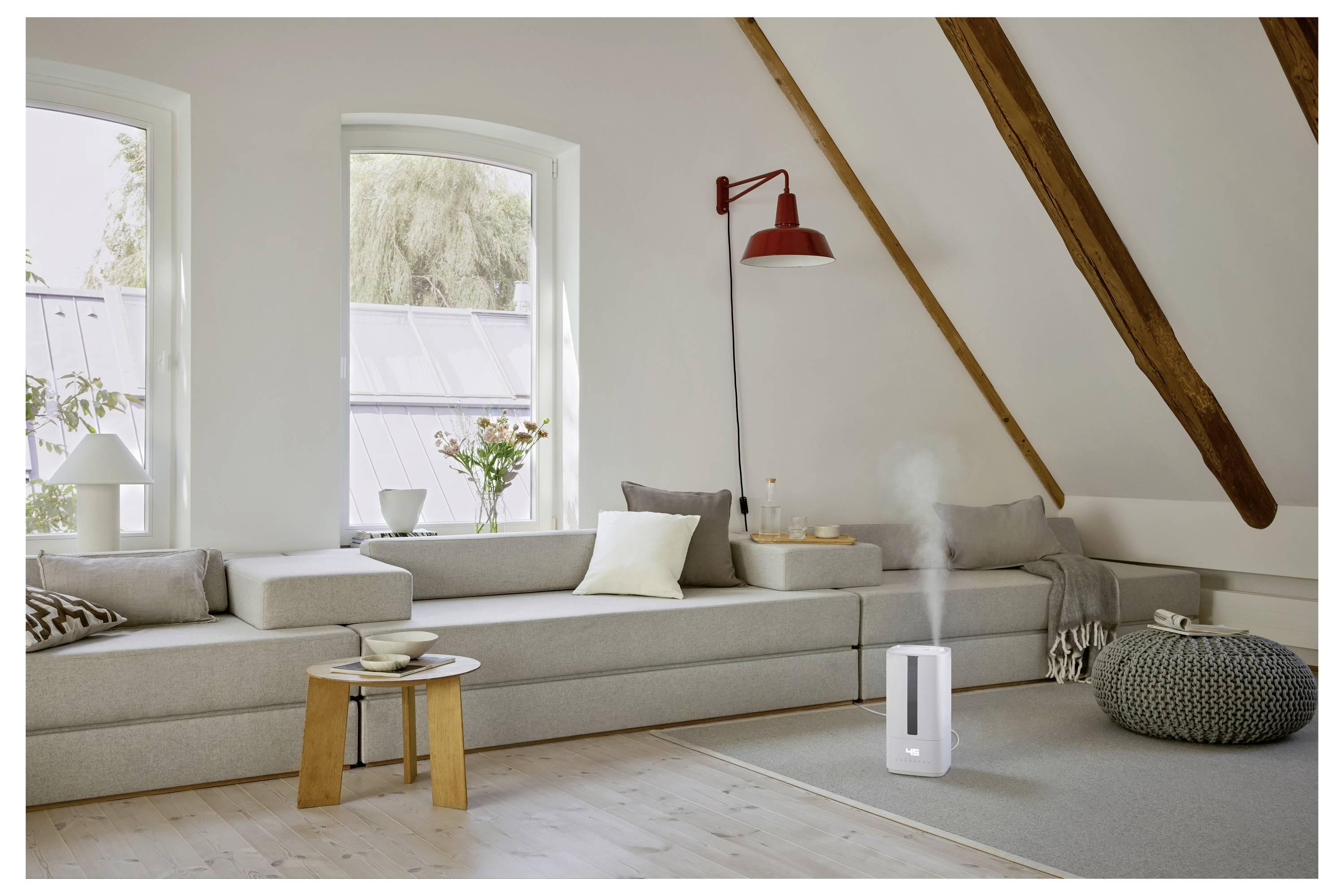 Ein gemütliches Dachgeschoss-Wohnzimmer mit einem langen grauen Sofa, einer roten Wandlampe, dekorativen Kissen und einem Luftbefeuchter, der Wasserdampf auf einem Teppich abgibt.