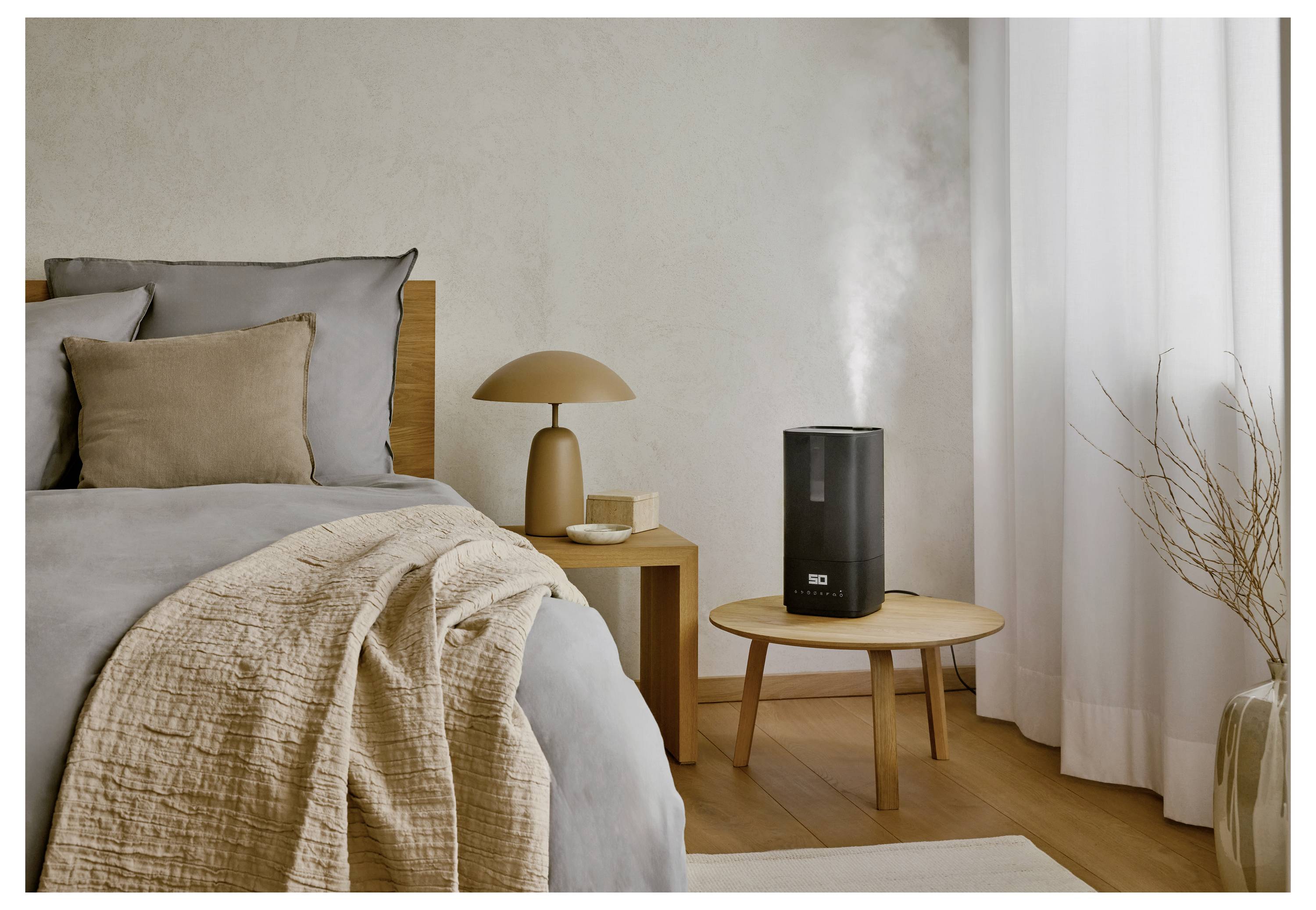 Ein modernes Schlafzimmer zeigt ein Bett mit beige-grauer Bettwäsche. Ein kleiner Holztisch trägt einen schwarzen Luftbefeuchter, der Nebel ausströmt, und eine Lampe.