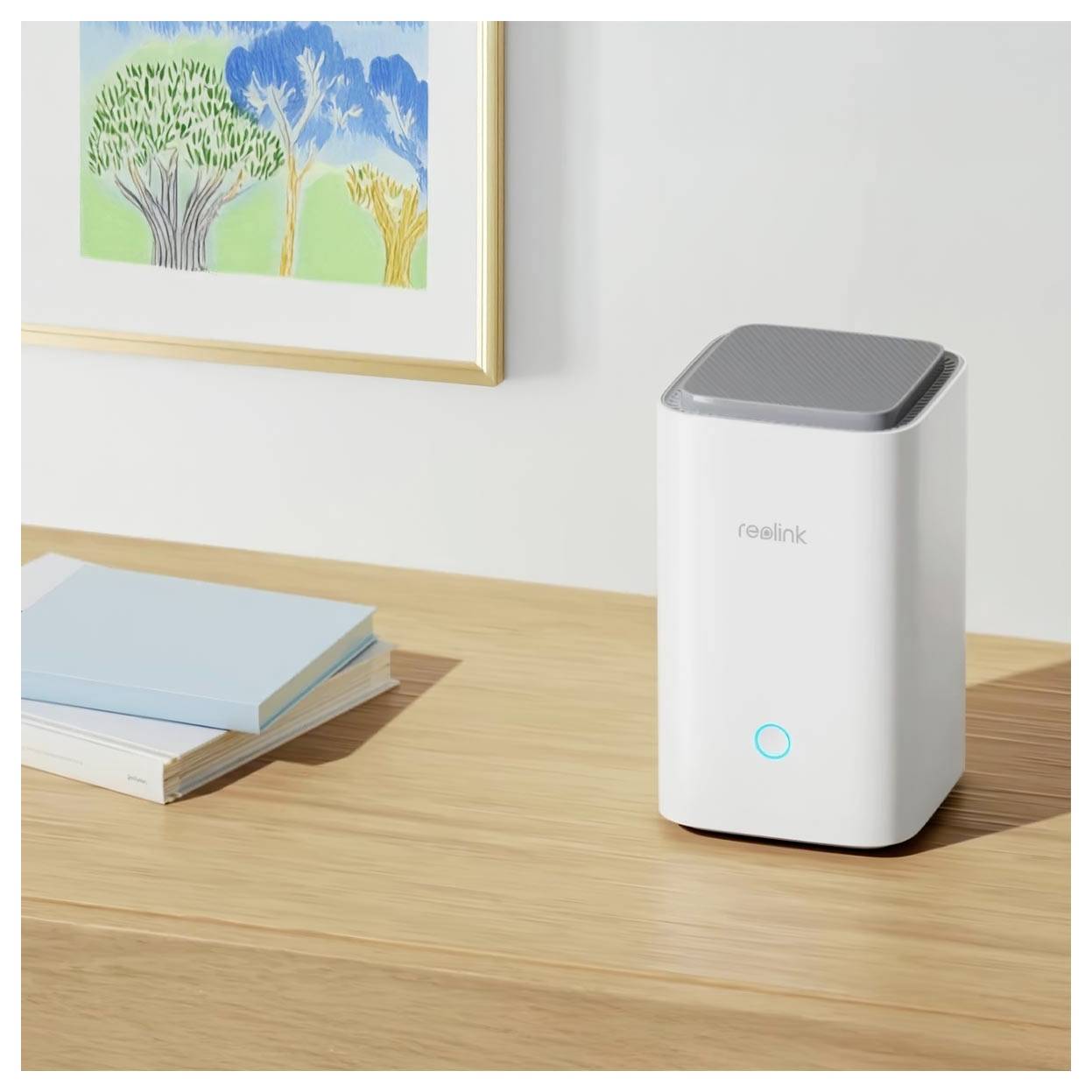 Ein weißes, rechteckiges Smart-Home-Gerät mit einem leuchtenden blauen Kreis sitzt auf einer Holzoberfläche neben zwei Büchern und einem Kunstwerk an der Wand.