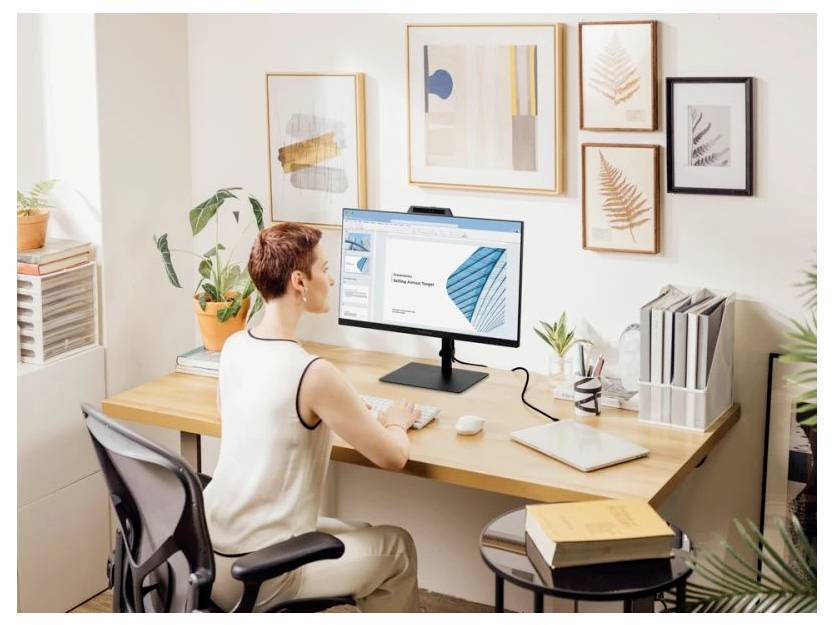 Eine Person, die an einem Schreibtisch sitzt und an einem Computer arbeitet. Der Monitor zeigt ein Dokument mit abstrakten Formen. Der Raum ist mit Pflanzen und gerahmten Kunstwerken dekoriert.