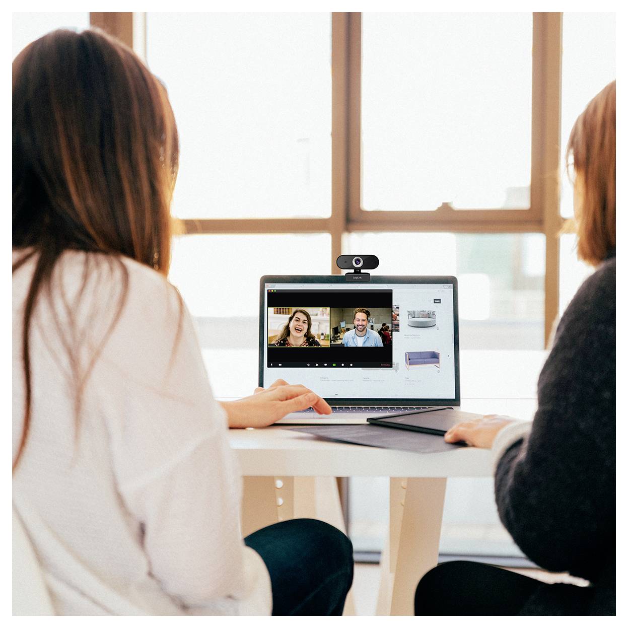 Zwei Personen, die an einem Tisch sitzen und an einem Videoanruf auf einem Laptop teilnehmen. Der Bildschirm zeigt einen Mann und eine Frau, die miteinander sprechen. Helles natürliches Licht.