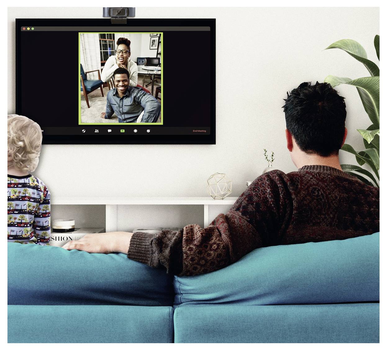 Eine Person und ein Kind sitzen auf einem Sofa und schauen einen Videoanruf auf einem Fernsehbildschirm, der zwei lächelnde Menschen in einer Hausumgebung zeigt.