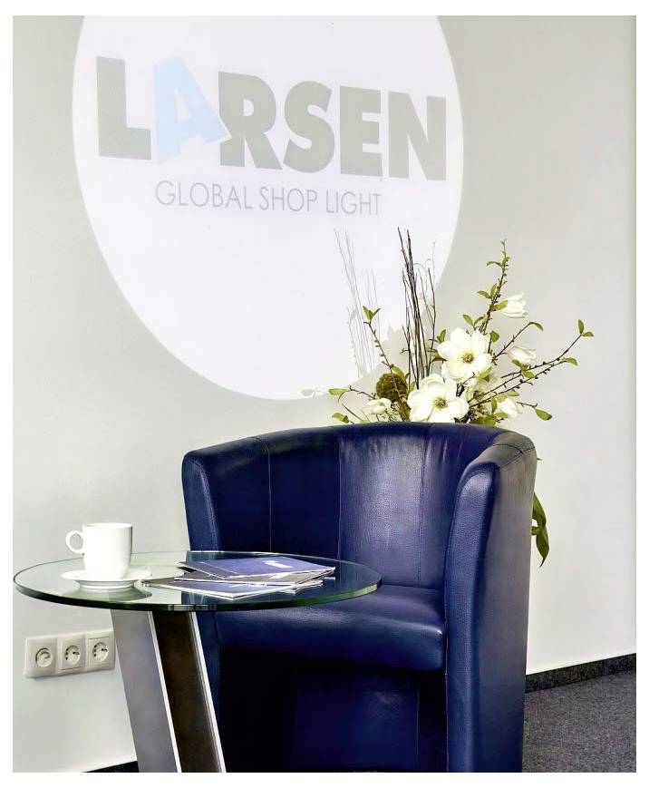 Ein blauer Ledersessel mit einer Blumenarrangement und einem kleinen Glastisch, auf dem eine Tasse und eine Zeitschrift stehen, vor einer Wand mit dem Logo "LARSEN GLOBAL SHOP LIGHT".