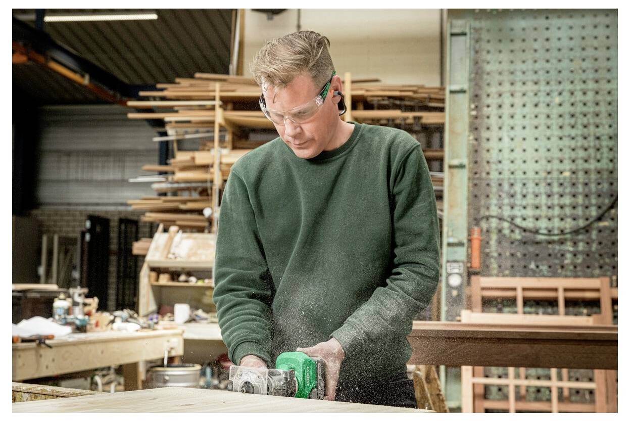 Eine Person mit Schutzbrille bedient ein Elektrowerkzeug in einer Holzwerkstatt, umgeben von Holzstapeln und Werkstattausrüstung.