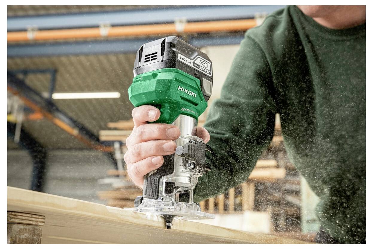 Eine Person benutzt einen grün-schwarzen elektrischen HandRouter auf einer Holzoberfläche in einer Werkstattumgebung, wodurch Sägespäne umherwirbeln.