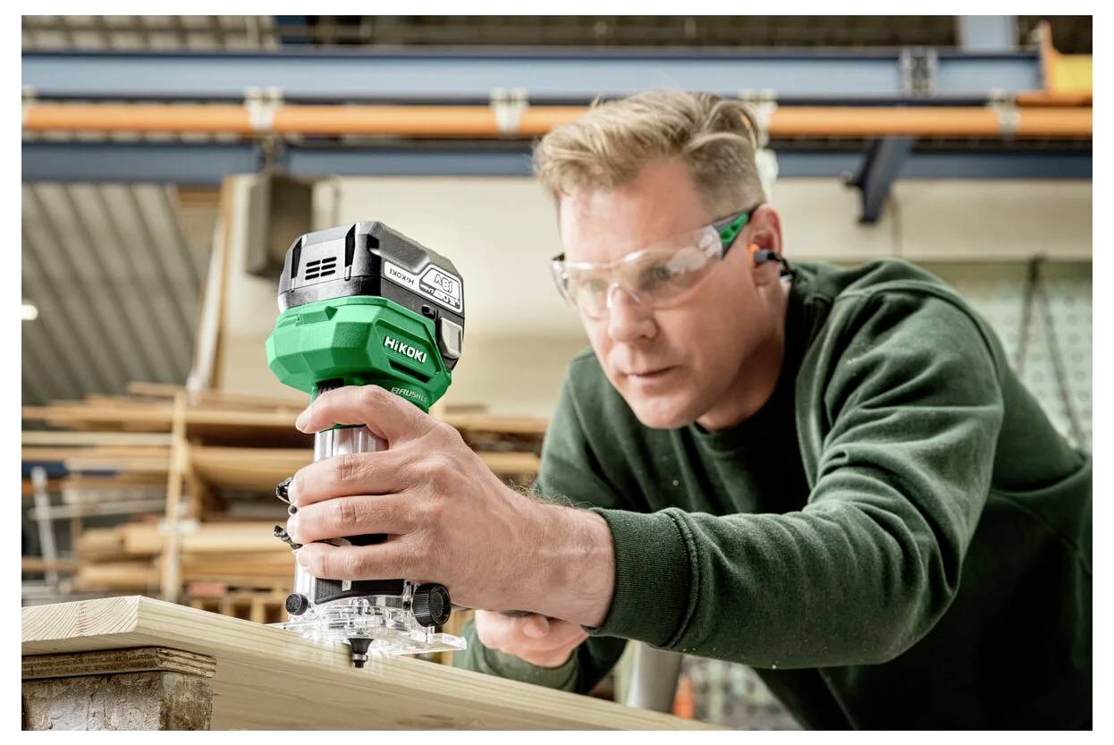 Eine Person mit Sicherheitsbrille bedient eine grün-schwarze Handoberfräse auf einem Holzbrett in einer Werkstattumgebung.