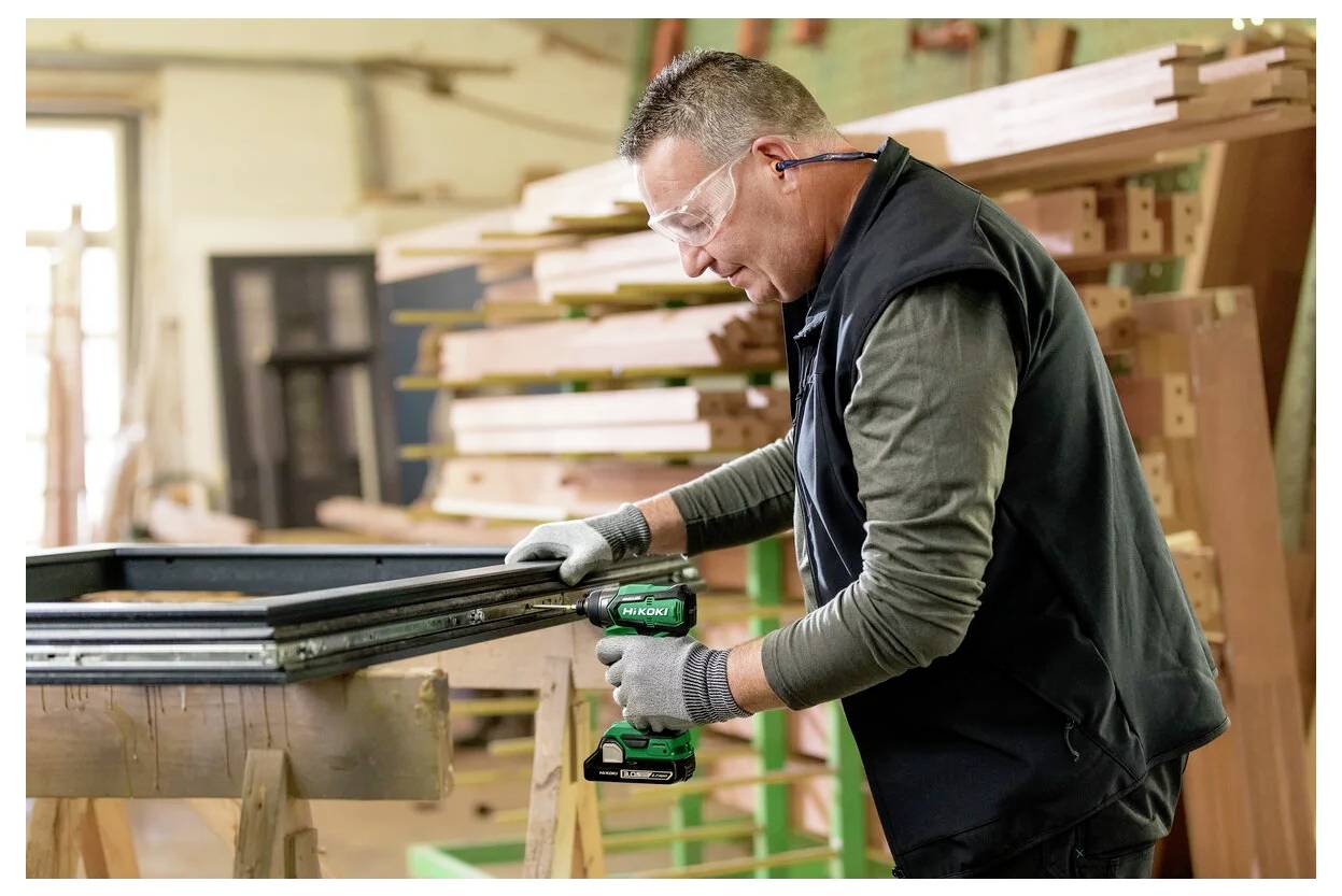 Ein Mann mit Sicherheitsbrille und Handschuhen verwendet einen Akkuschrauber, um einen Holzrahmen in einer Werkstatt voller Holzmaterialien zusammenzubauen.