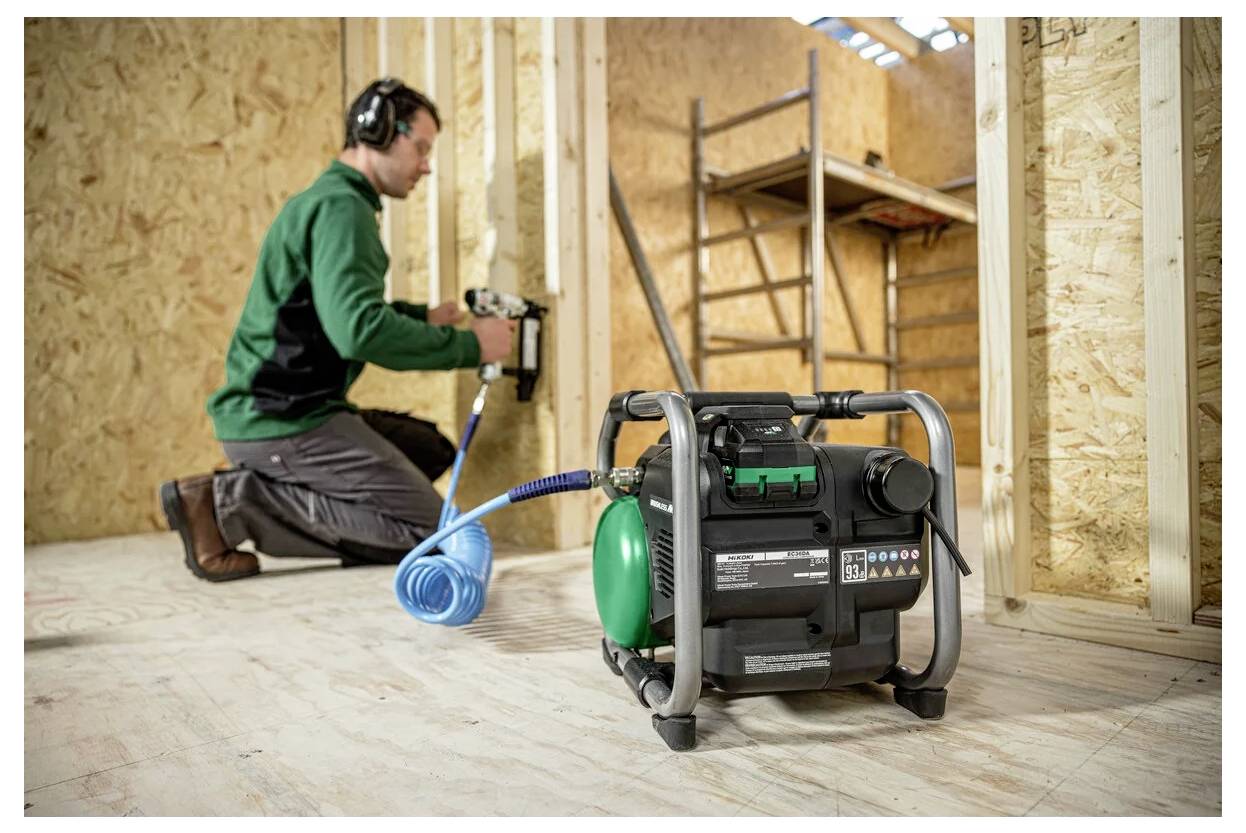 Eine Person mit Kopfhörern kniet auf einem Holzboden und bedient eine Nagelschussmaschine, die an einen grün-schwarzen Luftkompressor angeschlossen ist, in einer Baustellenumgebung.