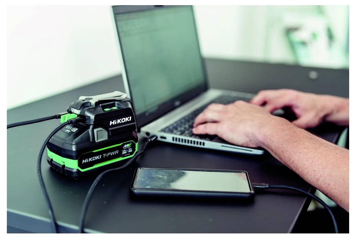 Eine Person tippt auf einem Laptop, während ein Hikoki-Akkuschrauber-Ladegerät daneben auf einem Tisch liegt, neben einem Smartphone.