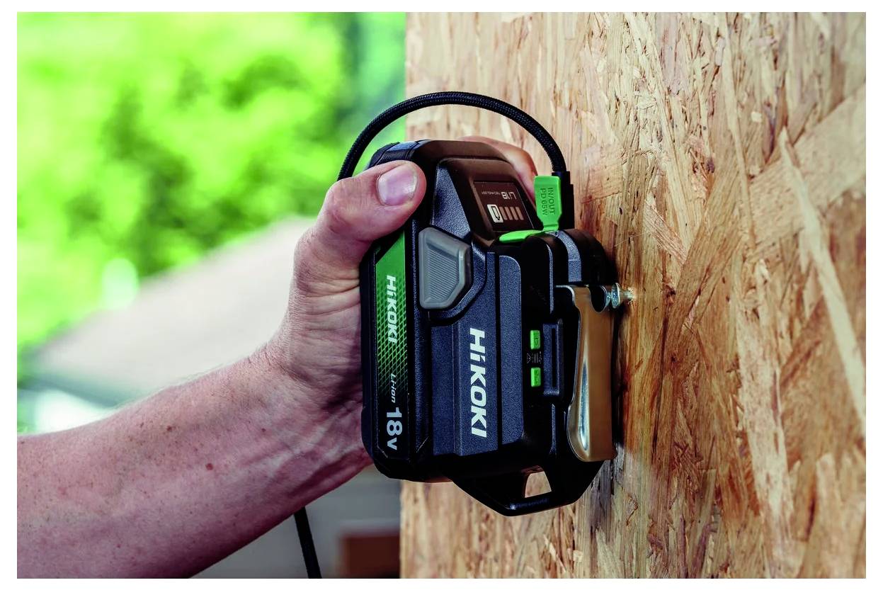 Hand, die ein grünes und schwarzes Hikoki 18V Elektrowerkzeug auf einer Sperrholzoberfläche im Freien verwendet, mit verschwommenem grünem Hintergrund.