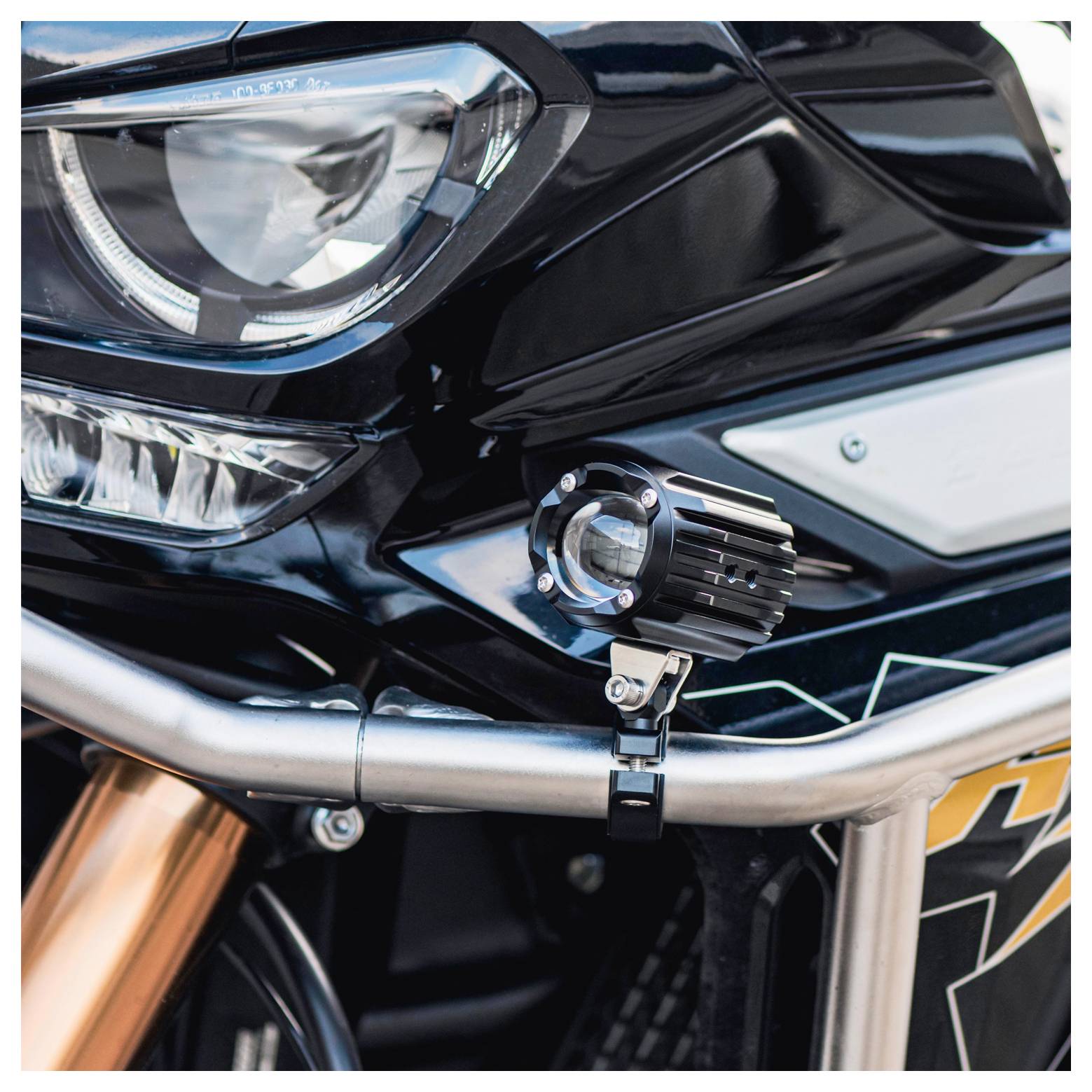 Eine Nahaufnahme der Frontpartie eines Motorrads, die eine Hauptscheinwerfer und eine zusätzlich montierte Hilfsleuchte am Rahmen des Fahrzeugs zeigt.