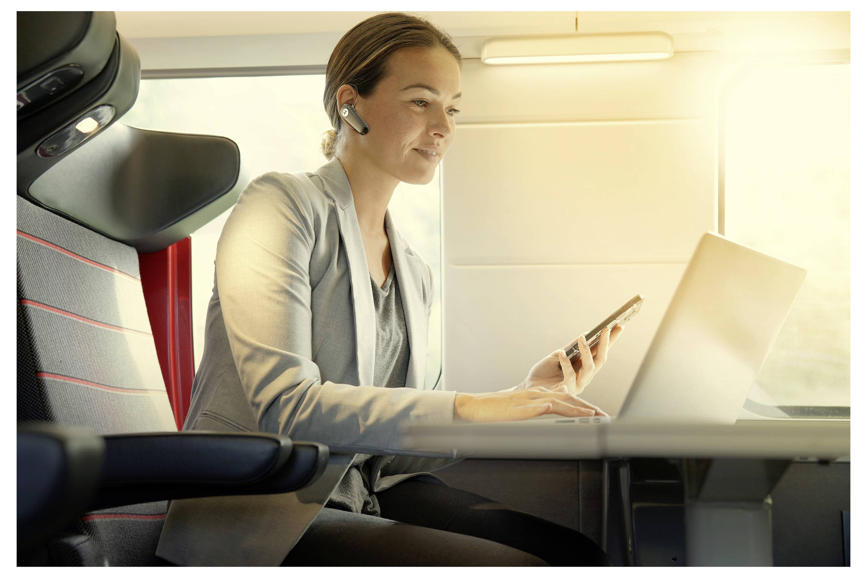 Eine Person in professioneller Kleidung arbeitet an einem Laptop, während sie ein Telefon hält und einen Headset trägt und bequem sitzt, was auf Multitasking hindeutet.