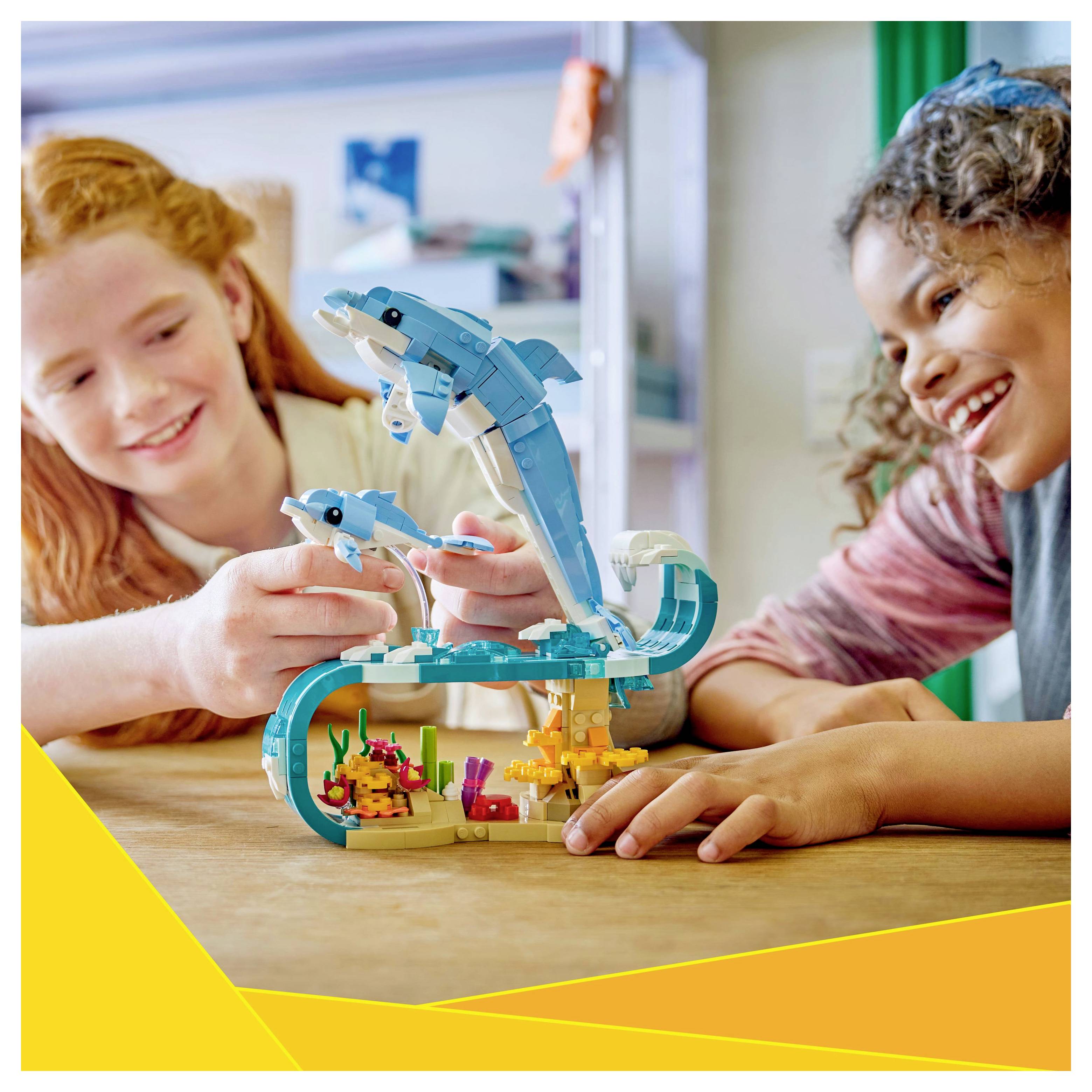 Zwei Kinder bauen ein buntes LEGO-Set mit Delfinen und Korallen und lächeln in einem hellen Raum.