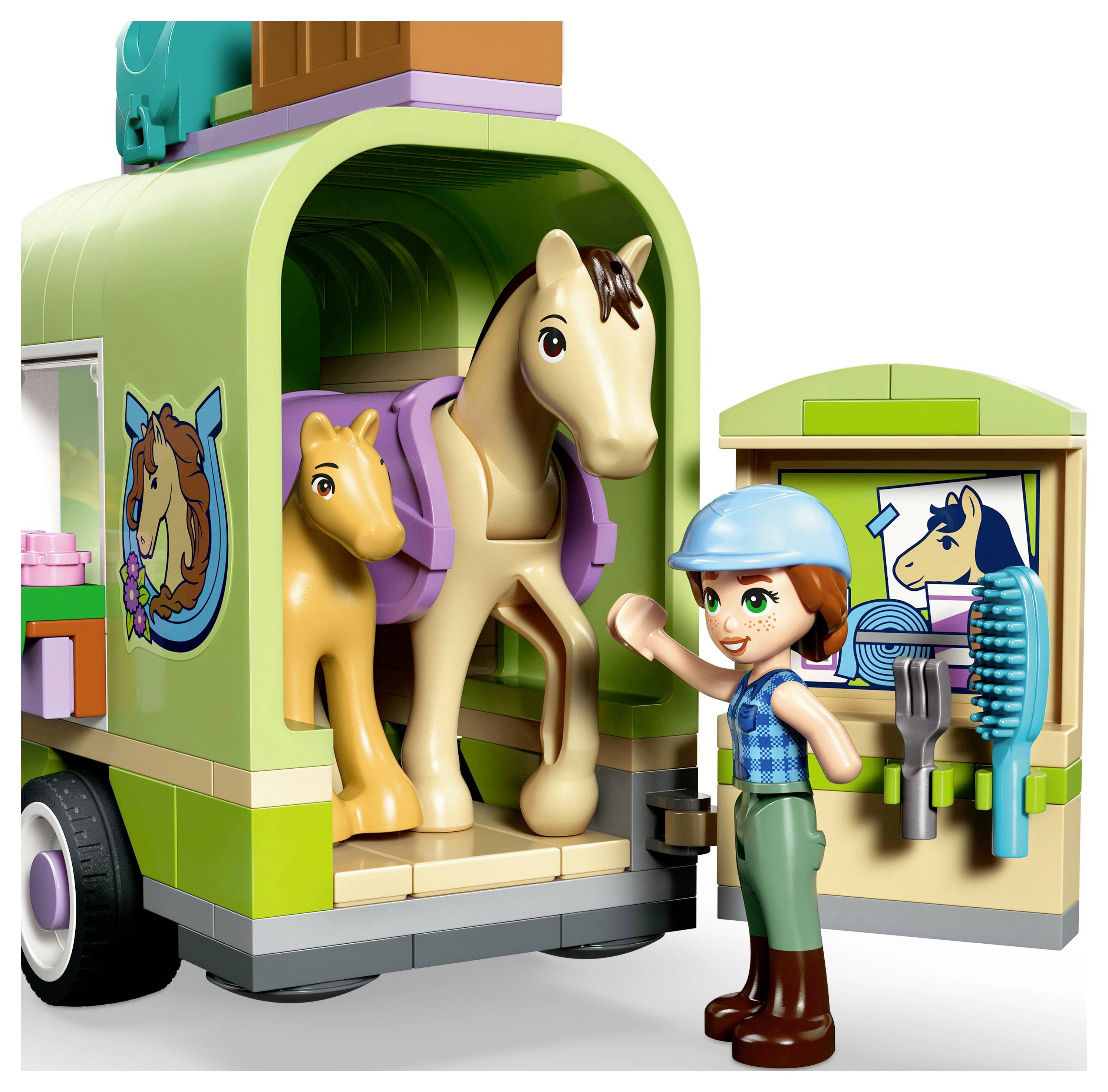 Ein Kind mit Helm winkt fröhlich neben einem Pferde-Anhänger für Spielzeugpferde, in dem zwei Spielzeugpferde stehen, und Pflegeutensilien, die an der offenen Tür ausgestellt sind.