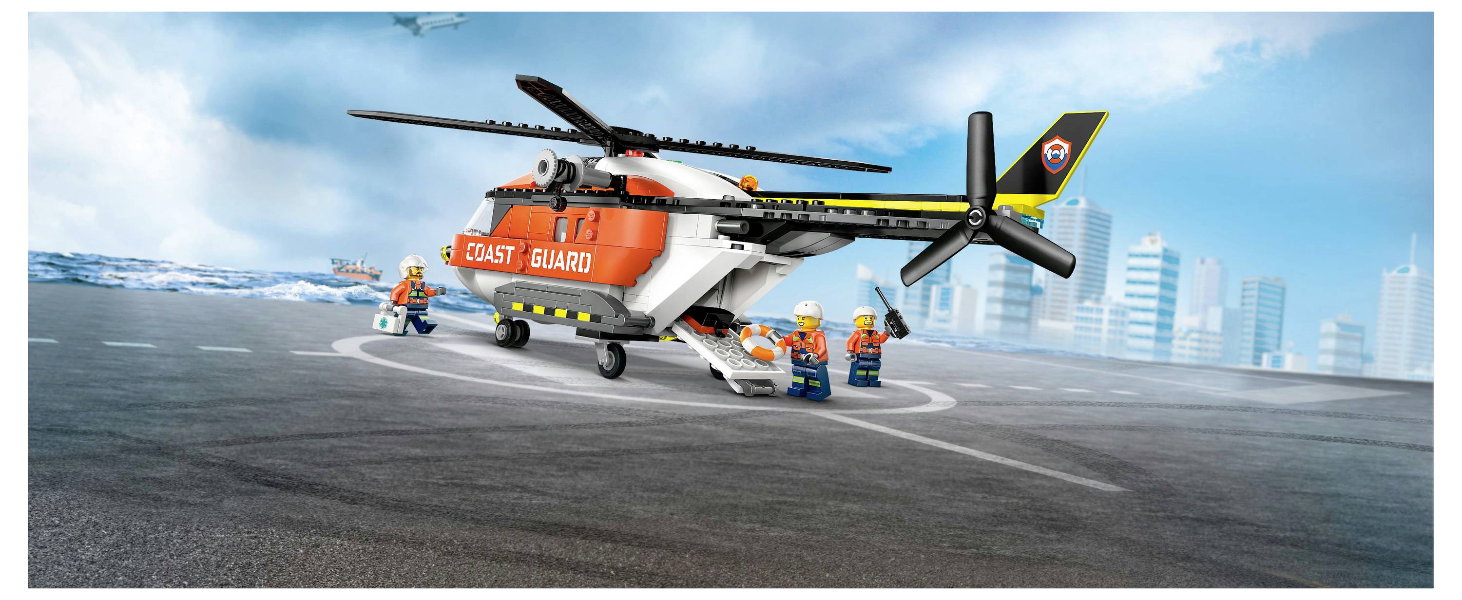 Ein Spielzeug-Küstenwachthubschrauber auf einer Startbahn, umgeben von drei Personen mit Sicherheitshelmem, mit einer Stadtsilhouette und dem Ozean im Hintergrund.