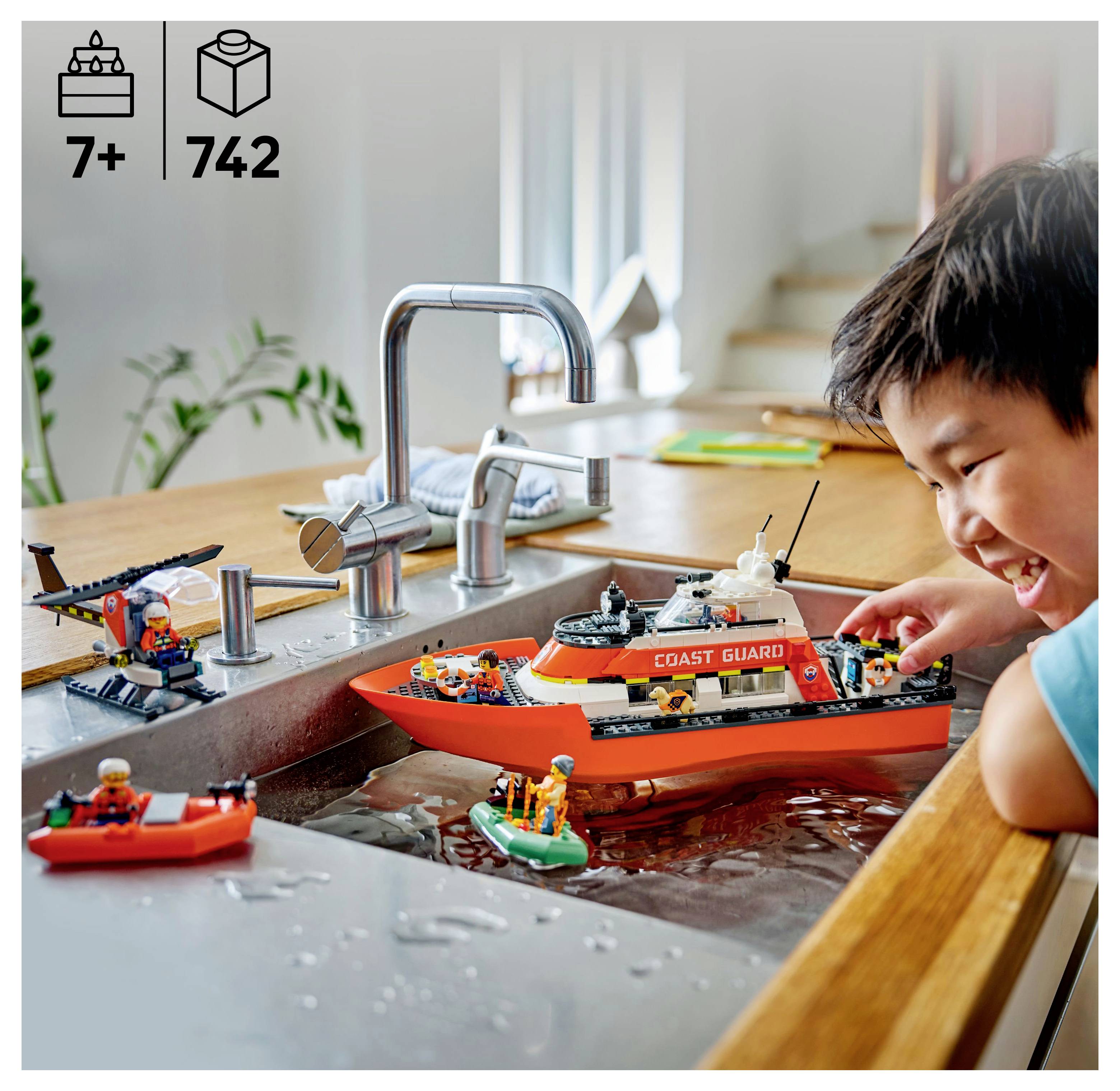 Ein Kind spielt freudig mit einem Küstenwache-LEGO-Set in einem mit Wasser gefüllten Spülbecken. Das Set umfasst ein Boot und einen Hubschrauber.