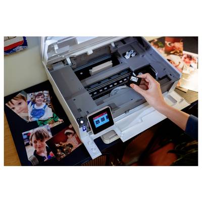 Eine Person ersetzt eine Tintenpatrone in einem Drucker, umgeben von gedruckten Fotos von Kindern, was auf eine Druckeinrichtung zu Hause hindeutet.