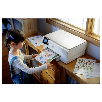 Ein Kind steht neben einem Drucker und holt farbenfrohe bedruckte Blätter mit verschiedenen Illustrationen heraus. Kunststoffmaterialien sind auf dem Tisch in der Nähe verstreut.