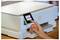 Eine Person interagiert mit dem Touchscreen-Interface eines modernen Druckers und wählt Optionen aus einem Menü aus, an einem Holzschreibtisch.