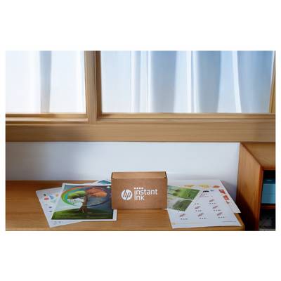 HP Instant Ink-Box auf einem Holztisch, umgeben von bunten Druckpapieren, neben einem Fenster mit weißen Gardinen.