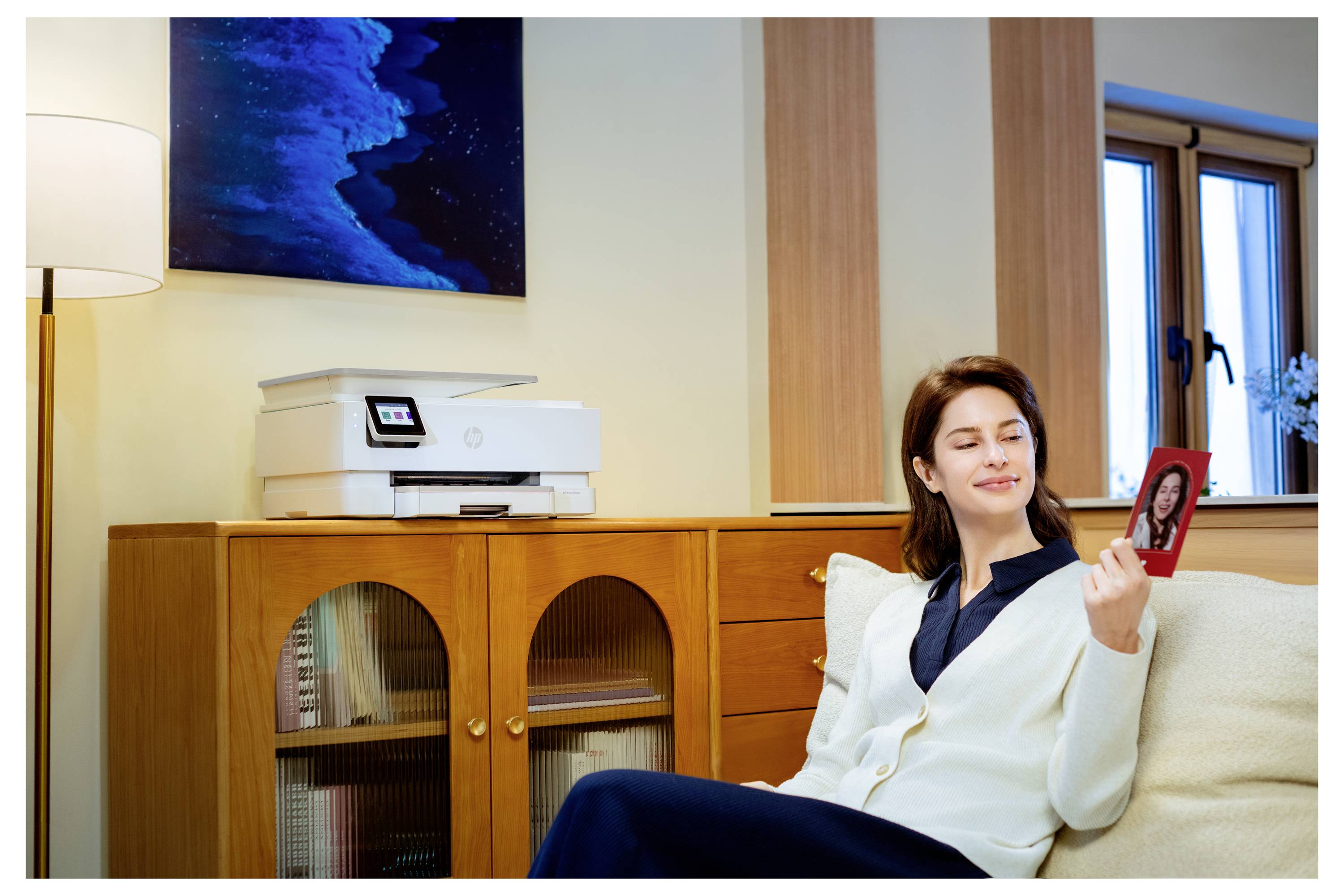 Eine Frau sitzt auf einem Sofa im Wohnzimmer und lächelt ein Foto an, das sie in der Hand hält. Ein Drucker steht im Hintergrund auf einem Schrank.