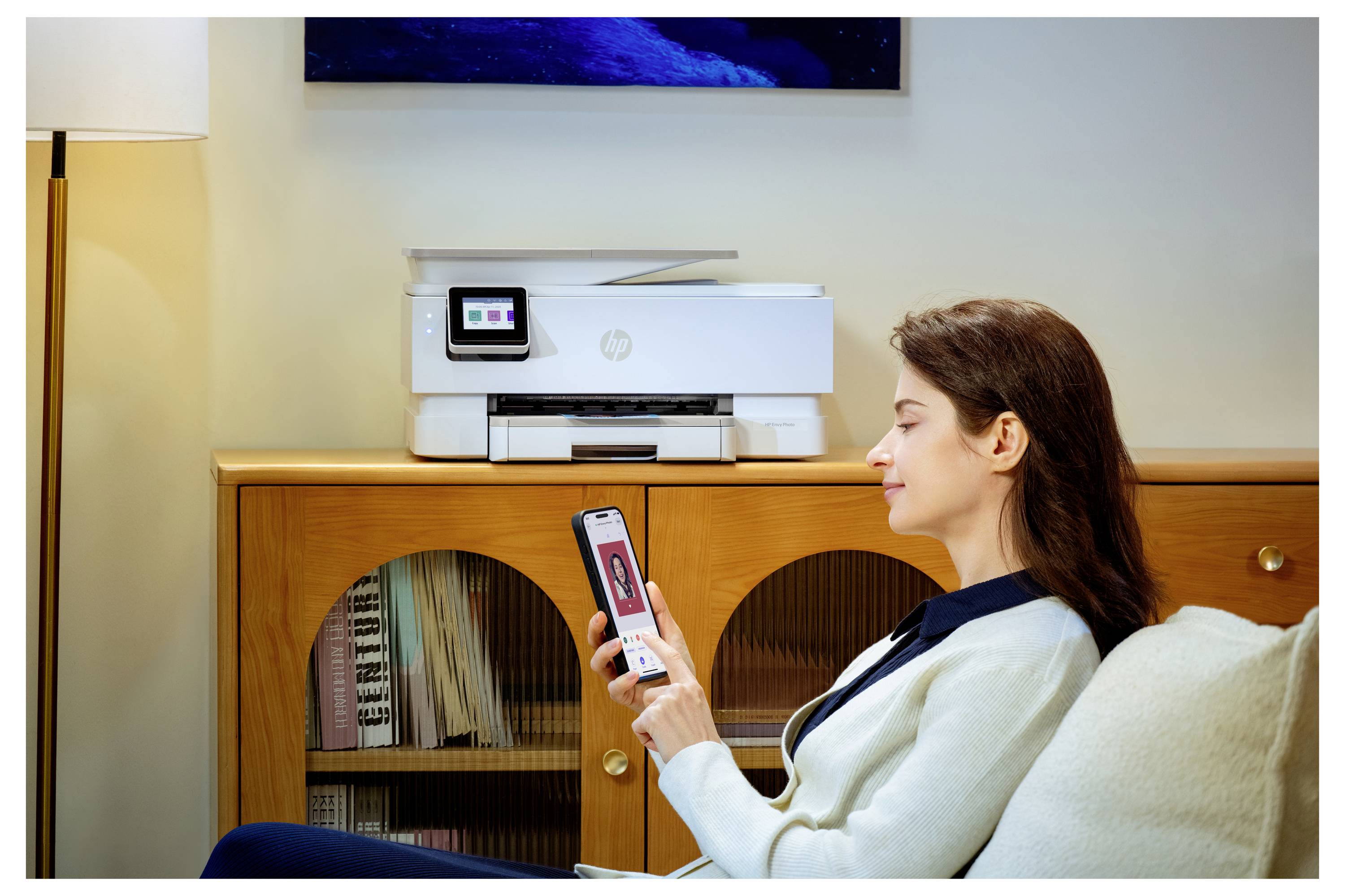 Eine Frau sitzt auf einem Sofa und verwendet ihr Smartphone, um drahtlos ein Foto auf einem Drucker zu drucken, der sich auf einem Holzschrank in der Nähe befindet.