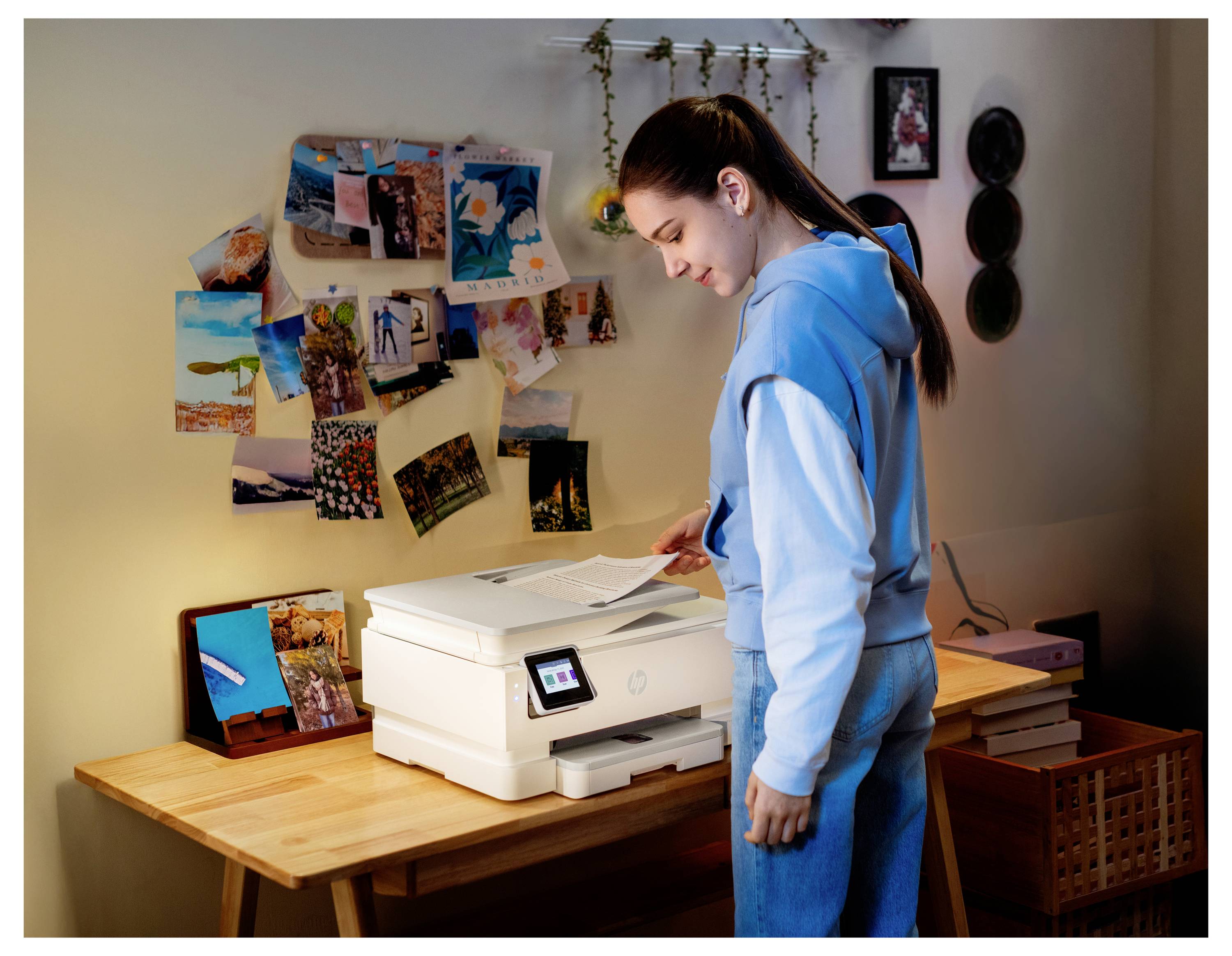 Eine Person in einem hellblauen Hoodie steht an einem Schreibtisch und druckt ein Dokument. Der Schreibtisch ist mit Fotos und kleinen Kunstwerken dekoriert.