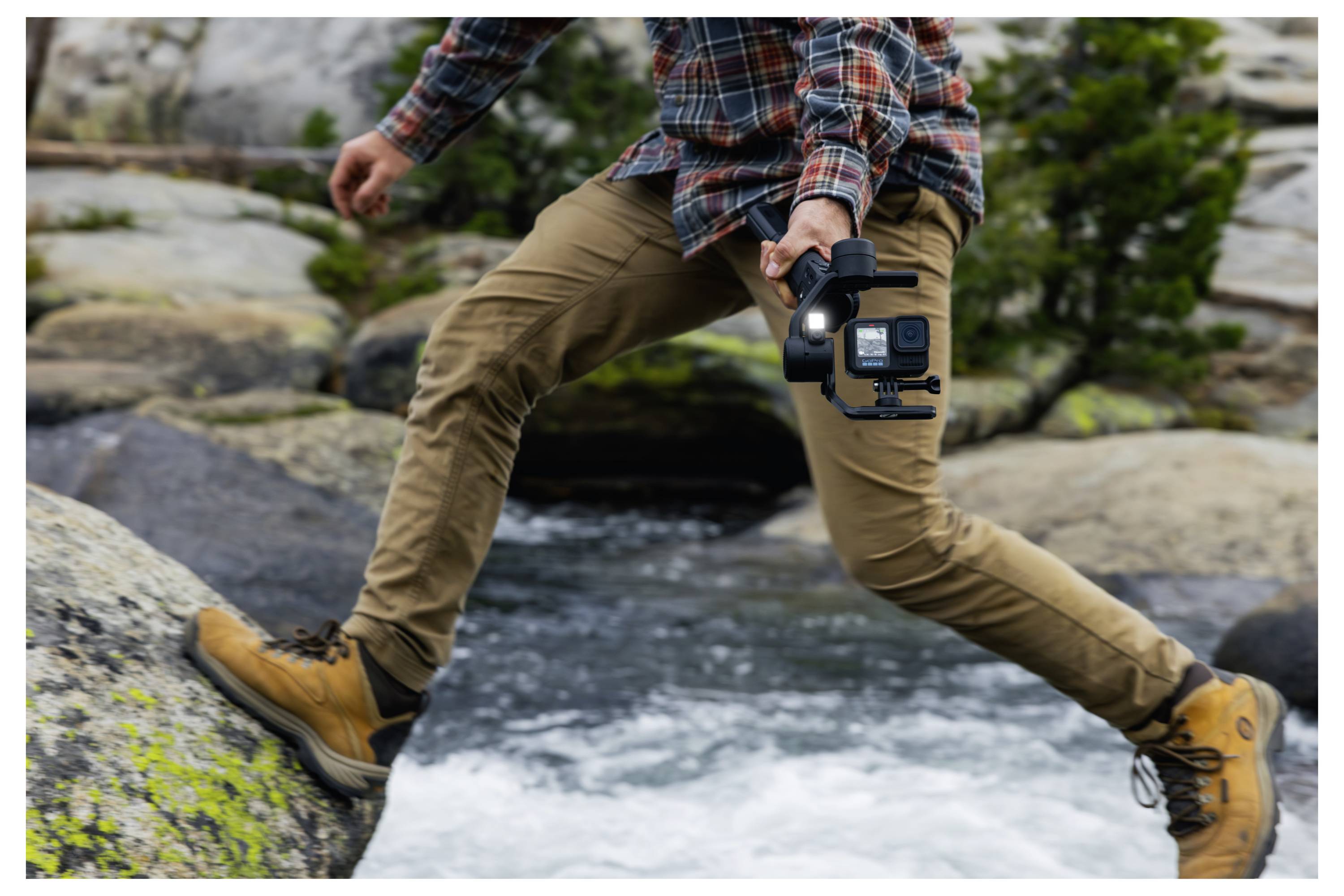 Eine Person in einem karierten Hemd und brauner Hose überquert einen felsigen Bach und hält eine Kamera auf einem Stabilisator, während sie Aufnahmen in der Natur macht.