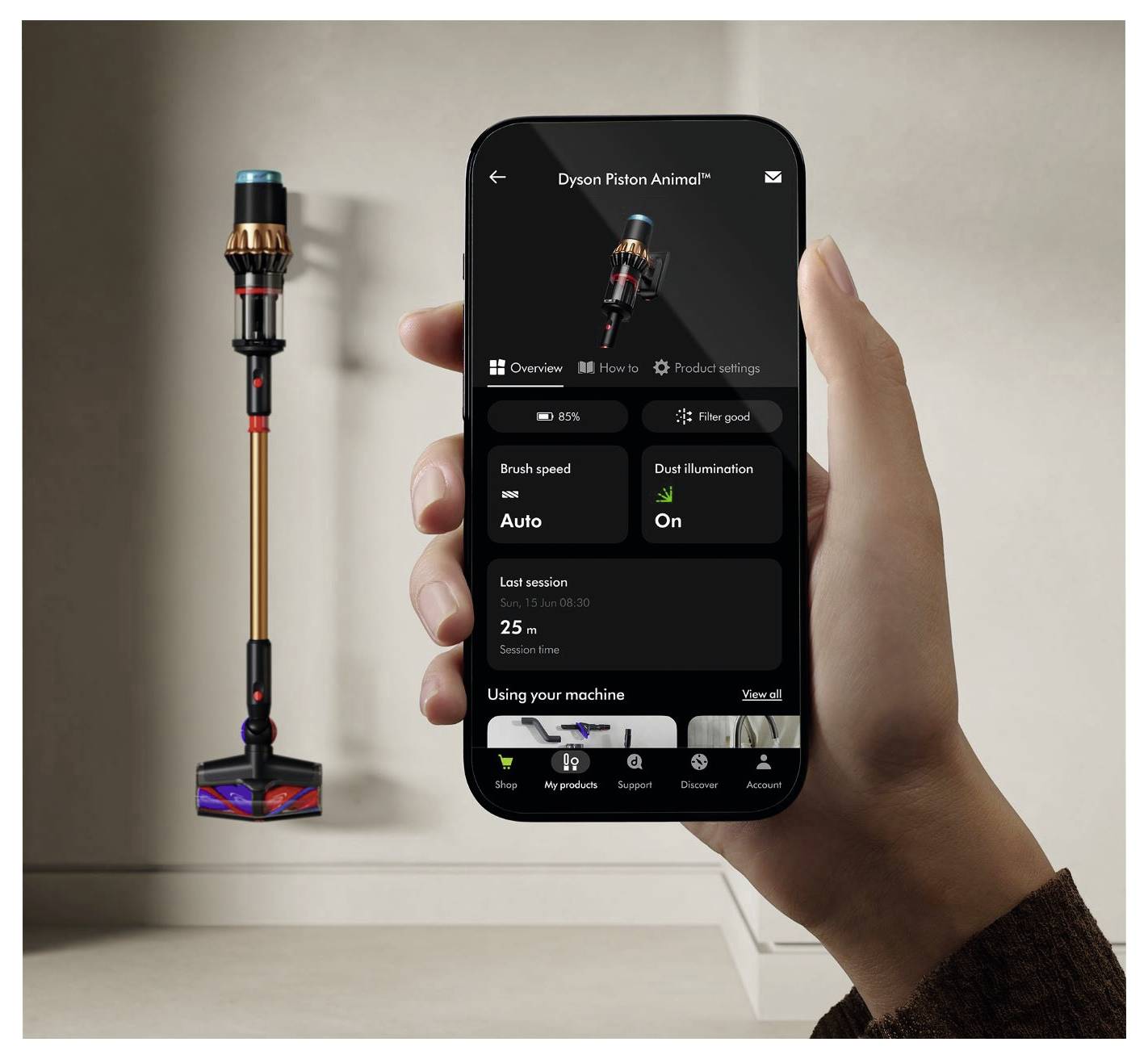 Eine Person hält ein Smartphone mit Einstellungen für einen Dyson-Staubsauger. Im Hintergrund ist ein kabelloser Staubsauger an einer Wand montiert.