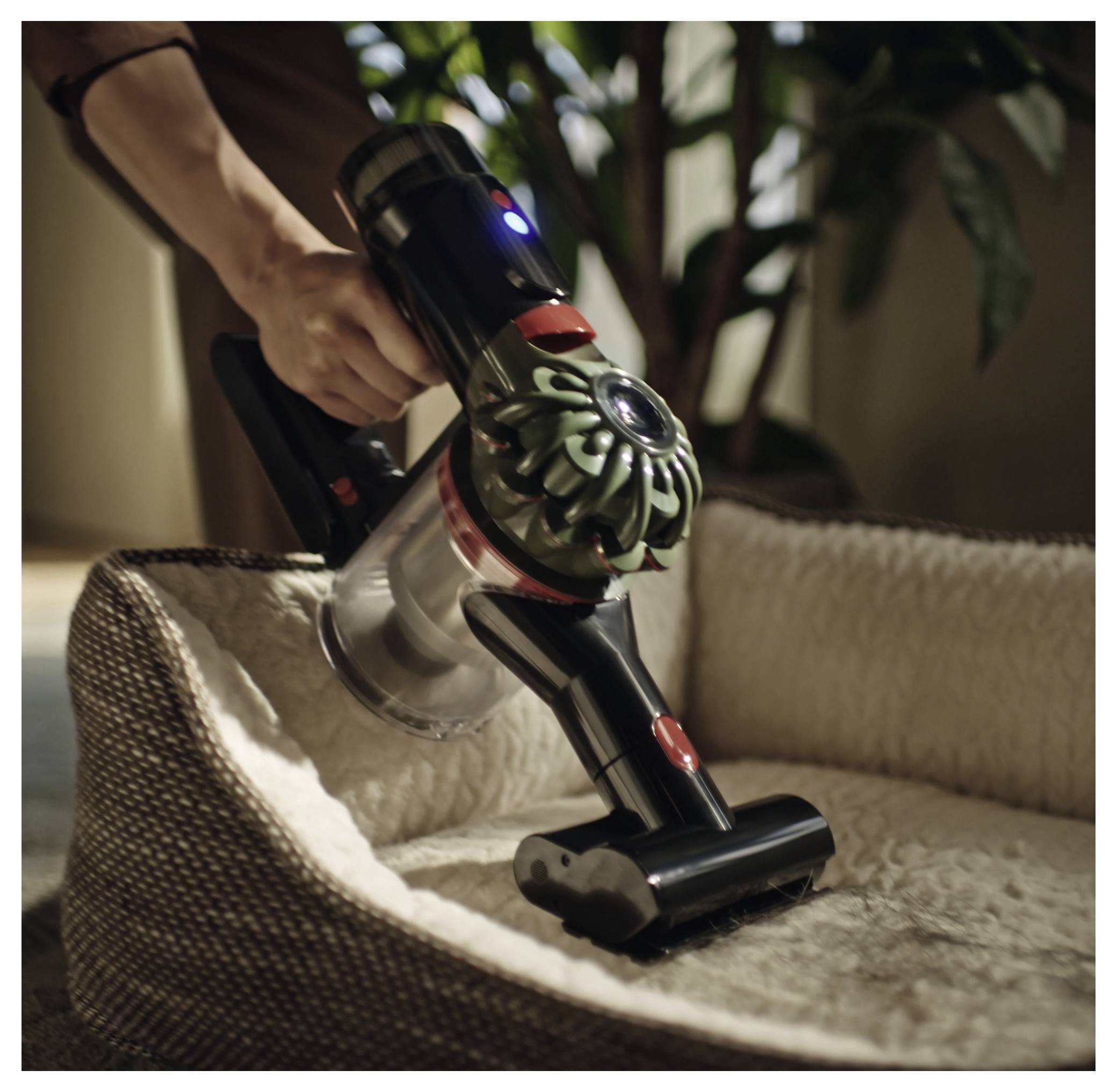 Eine Hand, die einen kabellosen Staubsauger hält und während des Reinigens ein beiges Haustierbett säubert, wobei Pflanzen im Hintergrund sichtbar sind.