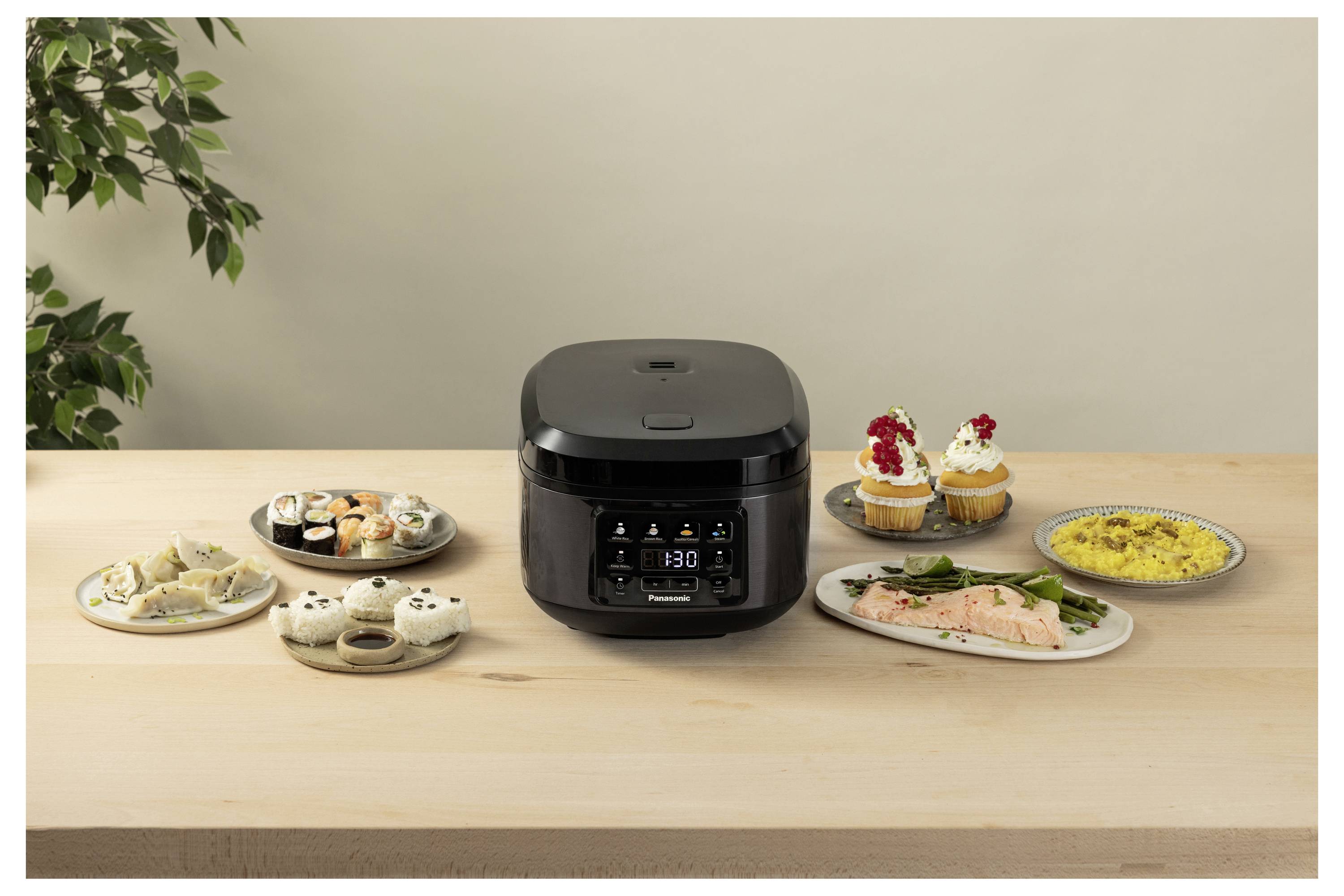 Ein schwarzer Multikocher steht auf einem Tisch, umgeben von verschiedenen Gerichten, darunter Sushi, Dumplings, Lachs, Cupcakes und Reis, mit einer Pflanze im Hintergrund.