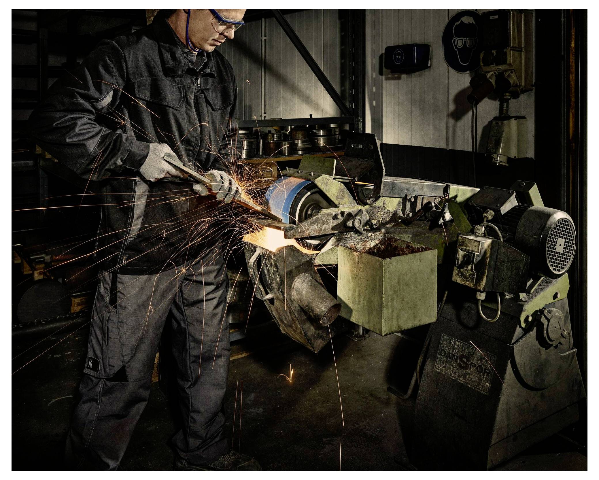 Ein Arbeiter in Schutzausrüstung schleift einen Metallzylinder an einer Industriemaschine, wobei Funken sprühen, in einer düster beleuchteten Werkstatt.