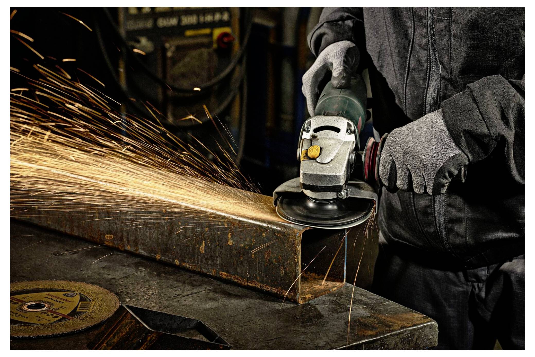 Eine Person mit Handschuhen bedient einen Winkelschleifer an einem Metallträger und erzeugt helle, umherfliegende Funken in einem industriellen Umfeld.