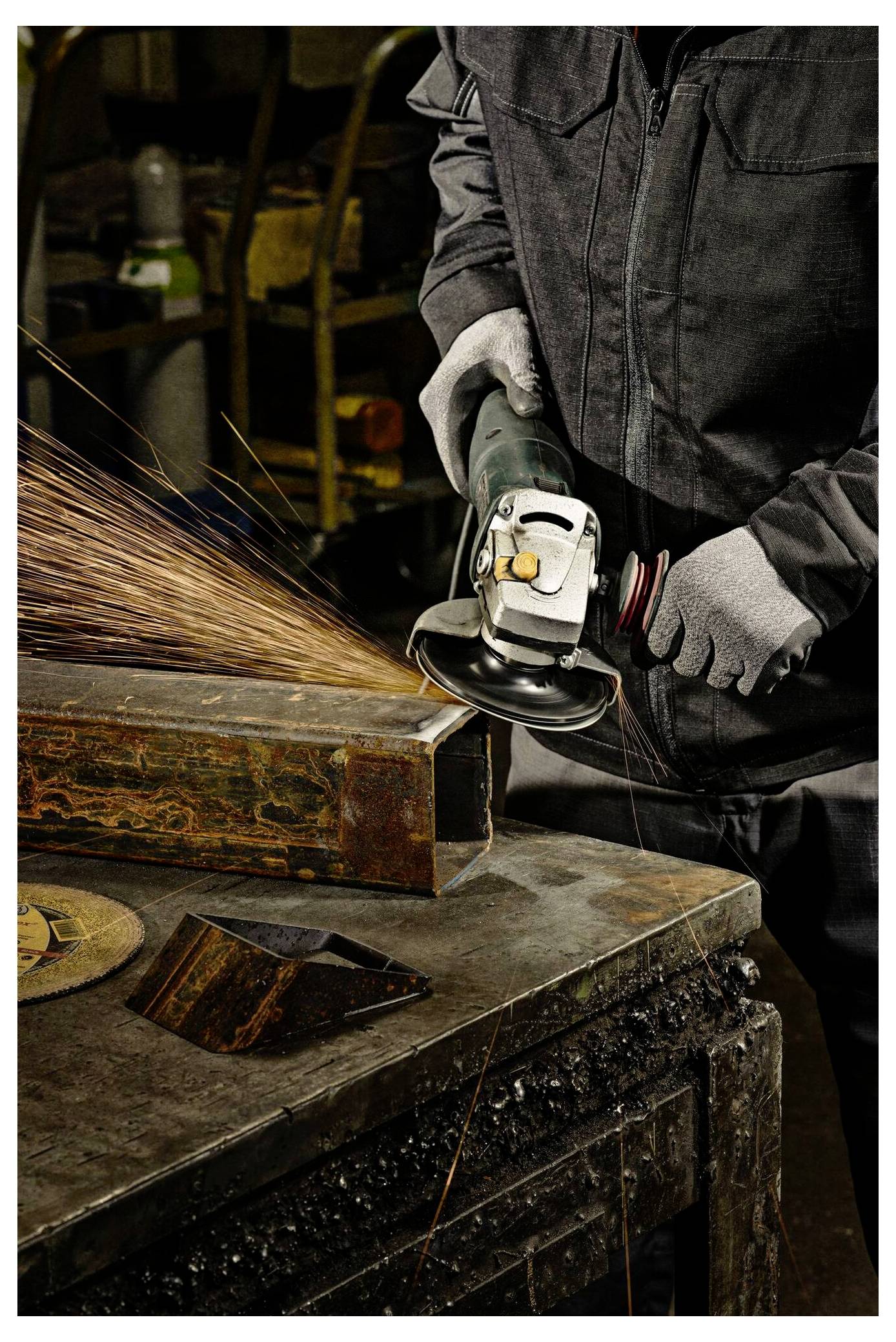 Eine Person mit Schutzausrüstung bearbeitet einen Metallträger mit einer Winkelschleifmaschine, wodurch Funken sprühen, in einer Industriewerkstatt.