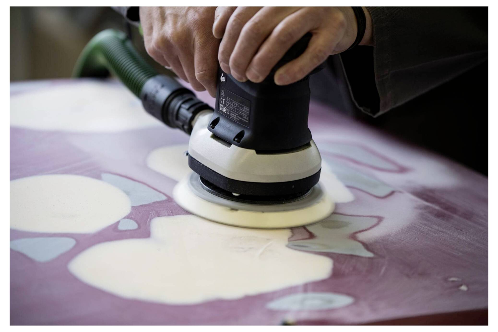 Eine Person benutzt einen elektrischen Schleifer auf einer Holzoberfläche, die mit Flecken von weißer und lila Farbe bedeckt ist, und konzentriert sich darauf, die Textur zu glätten.