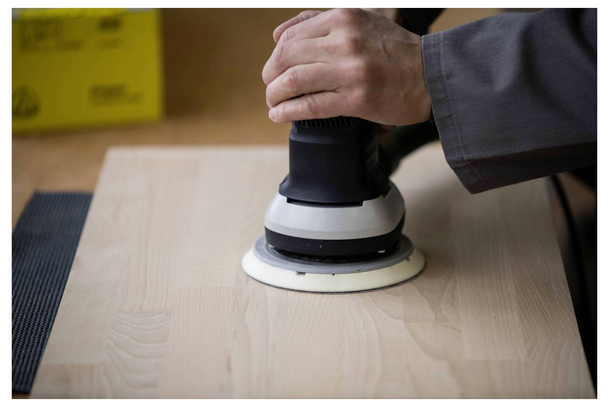 Hände, die einen elektrischen Schleifer verwenden, um eine helle Holzoberfläche zu glätten, mit einer gelben Box im Hintergrund.